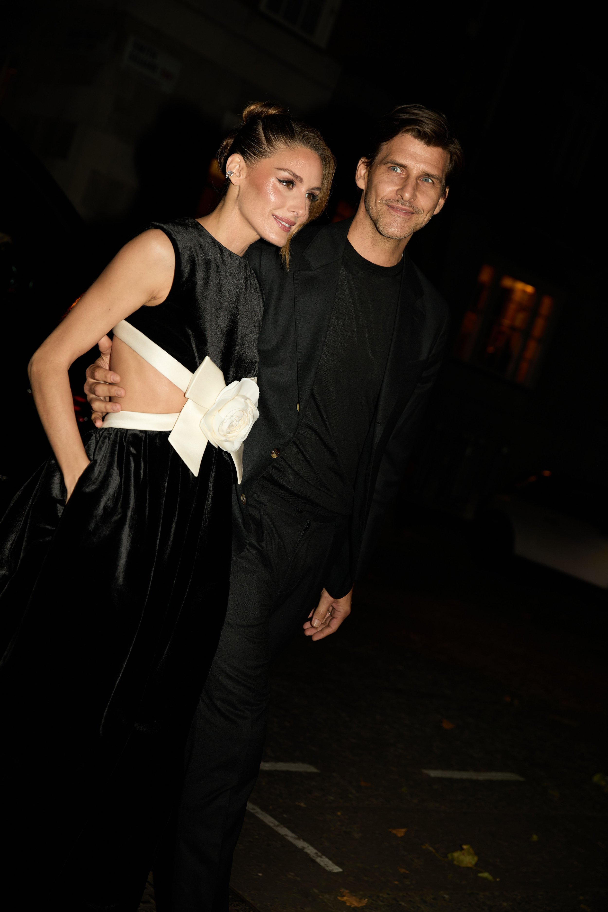 A woman and a man dressed in formal black attire posing together at night. The woman is wearing a black satin dress with a white flower detail and has her hair styled in an elegant updo. The man is wearing a black suit and shirt, with his arm around 