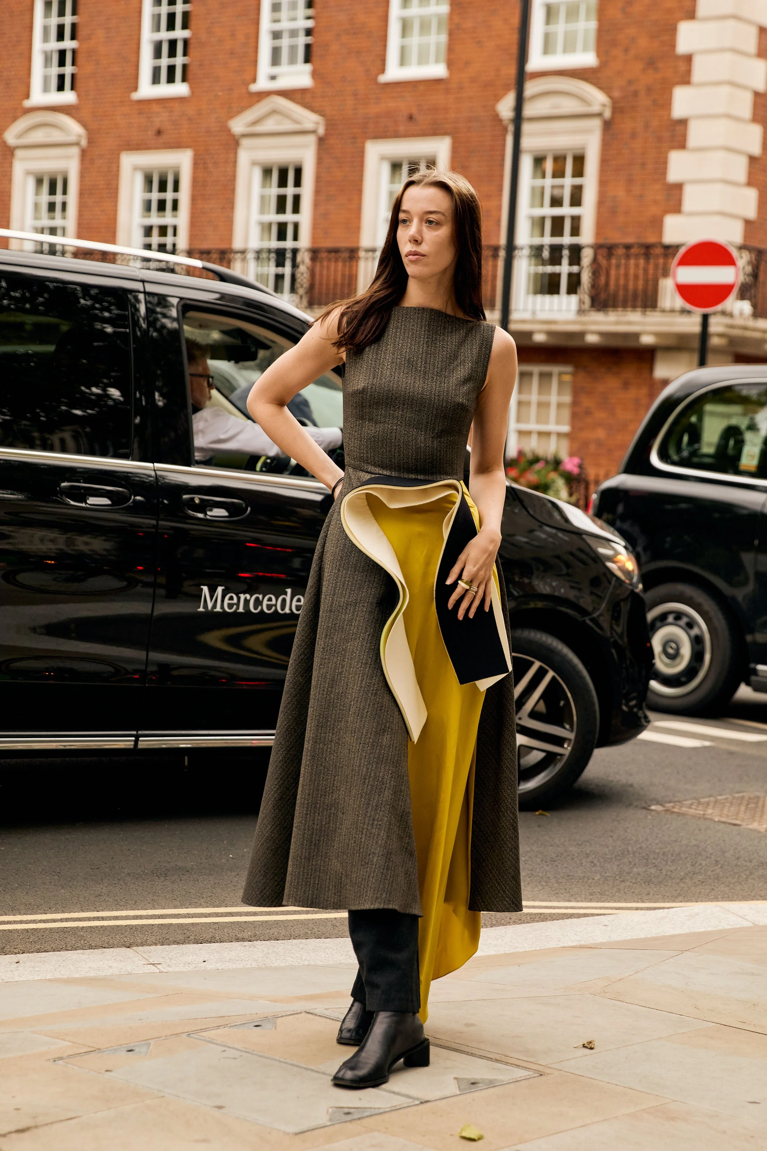 A woman in a grey sleeveless dress and black ankle boots standing on a city sidewalk in front of a black Mercedes vehicle, with a brick building and a no entry sign in the background.
