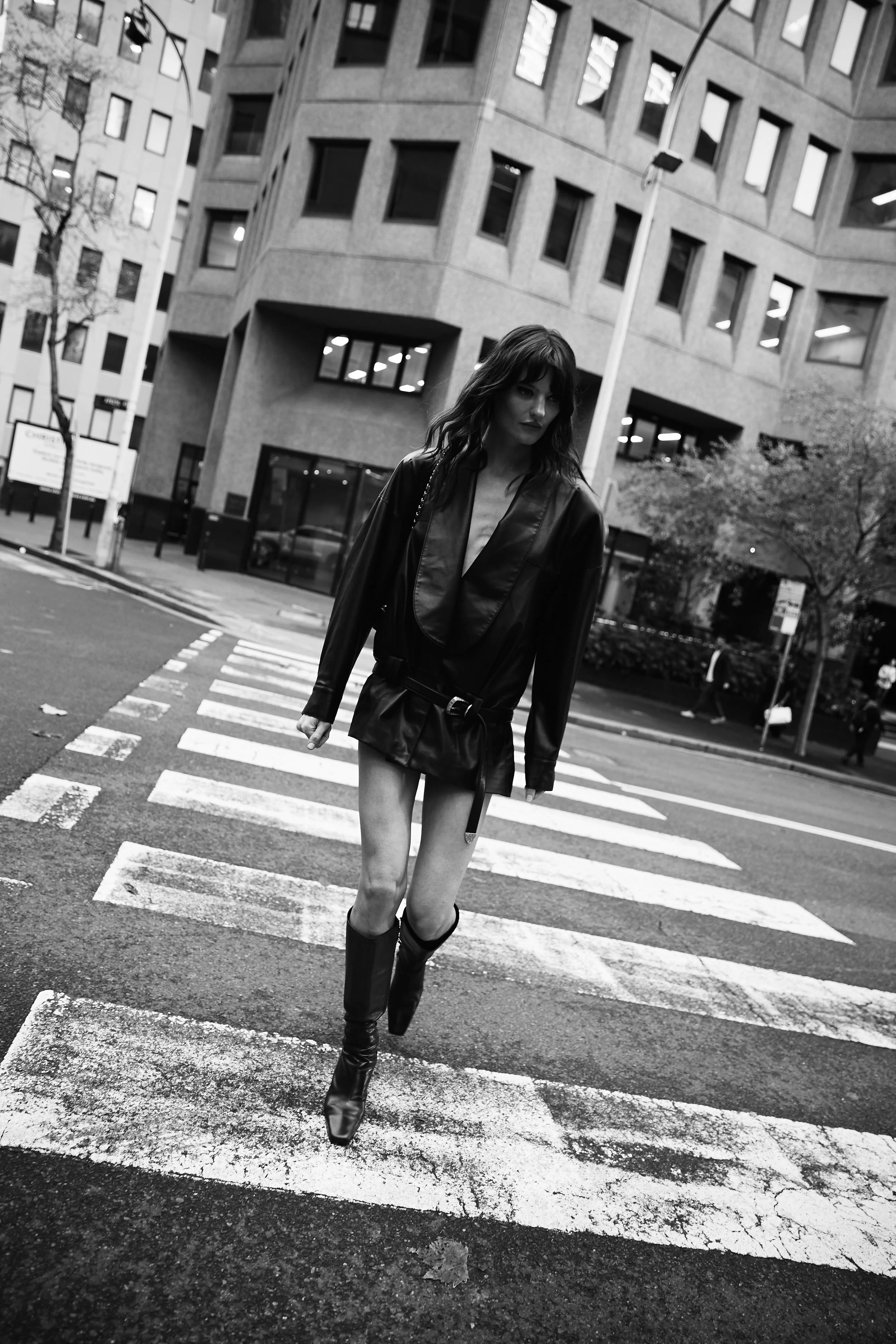 A woman with wavy hair and wearing a leather jacket and boots is walking across a crosswalk in an urban area.