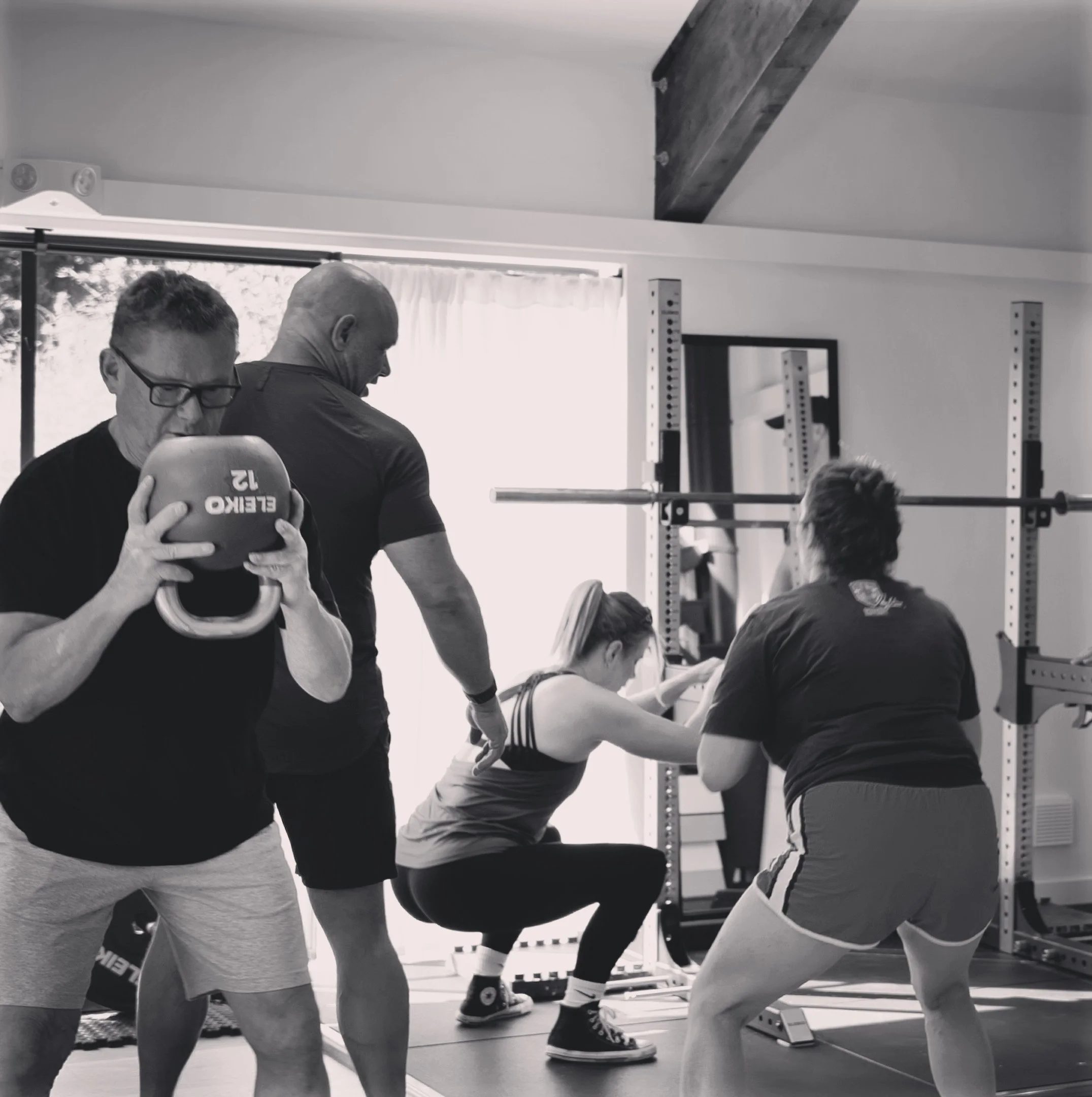 Three people in a gym, with two women and one man. One woman is kneeling, coaching the other woman who is lifting a barbell. The man is standing behind and slightly above them, observing. The scene is in black and white.
