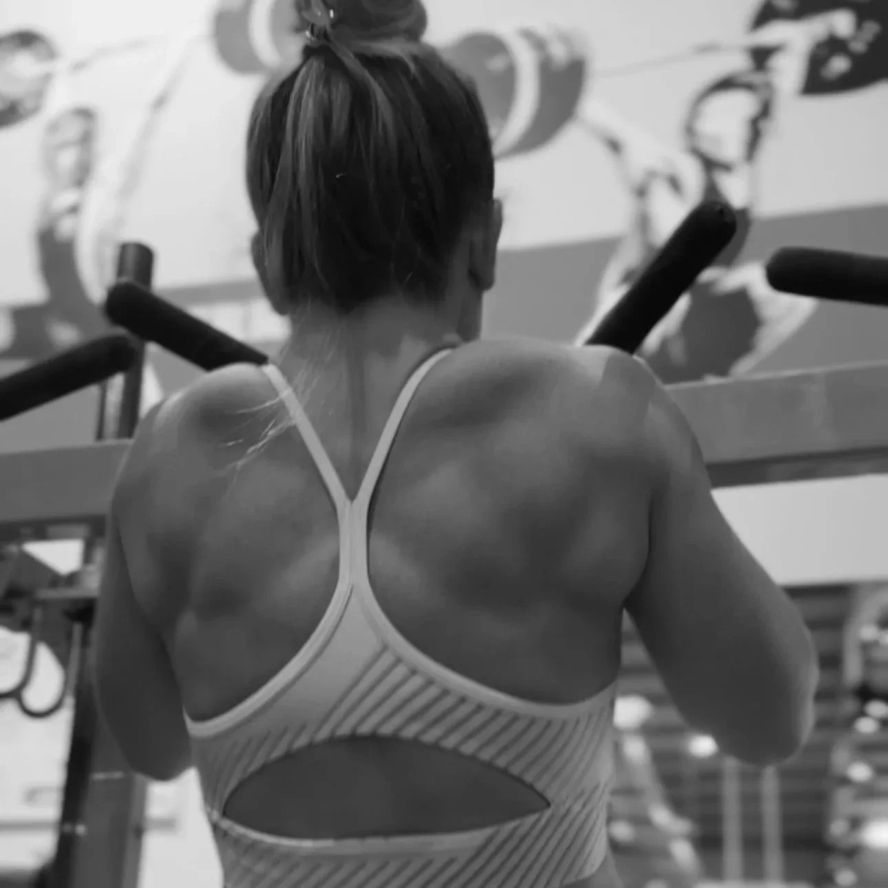Back view of a woman in workout attire exercising in a gym, with weightlifting equipment and a mural in the background.
