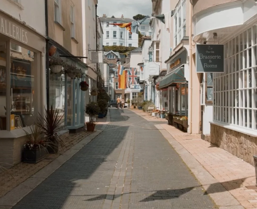 A narrow European street with shops, potted plants, colorful flags hanging across, and a handmade Union Jack banner in the background.