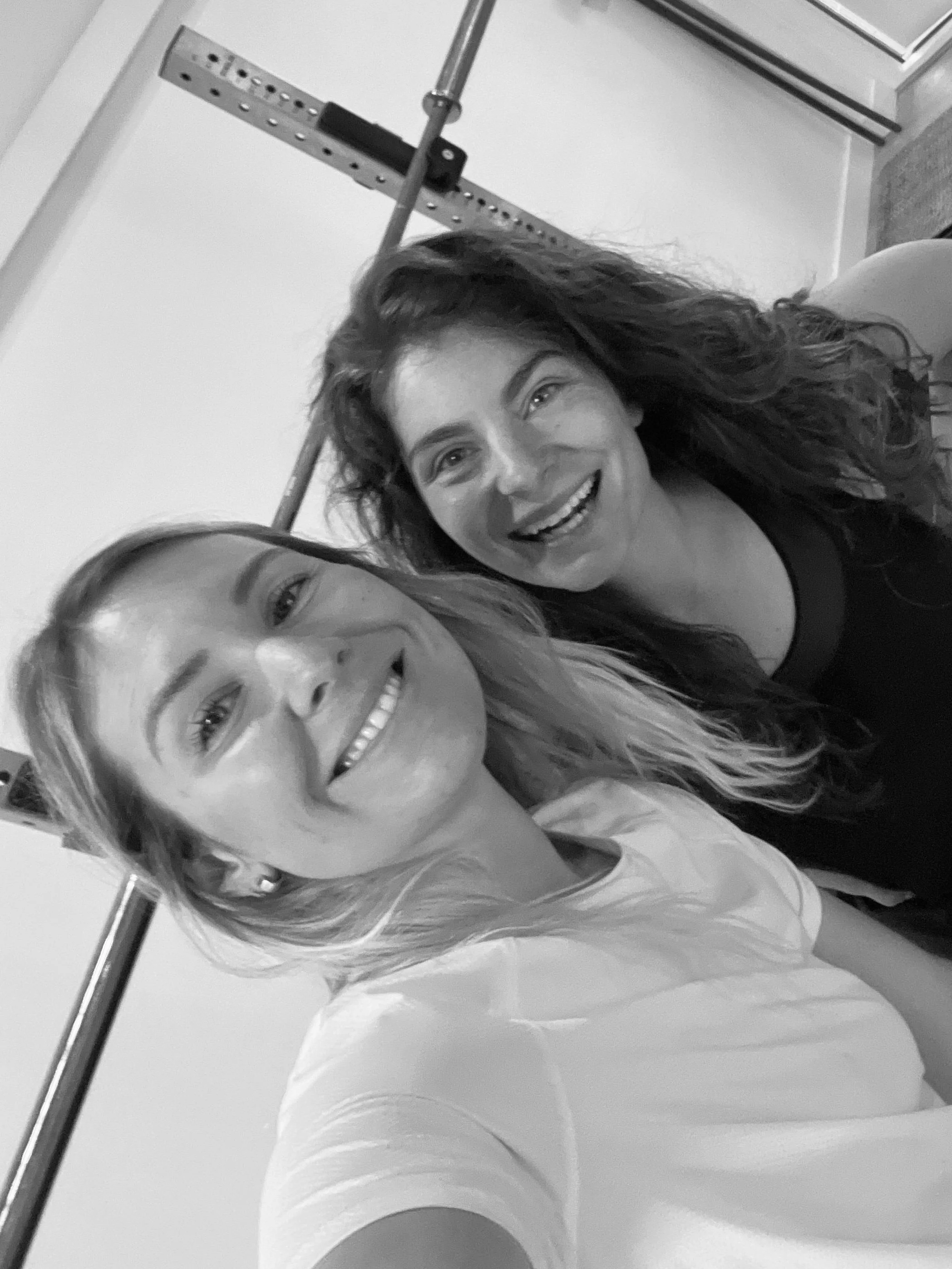 Three people in a gym, with two women and one man. One woman is kneeling, coaching the other woman who is lifting a barbell. The man is standing behind and slightly above them, observing. The scene is in black and white.