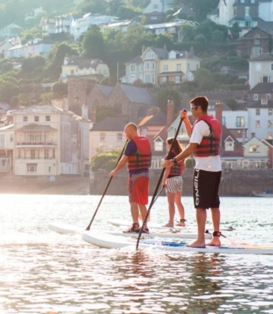 Three people paddleboarding on a body of water at sunset, with houses on a hillside in the background.