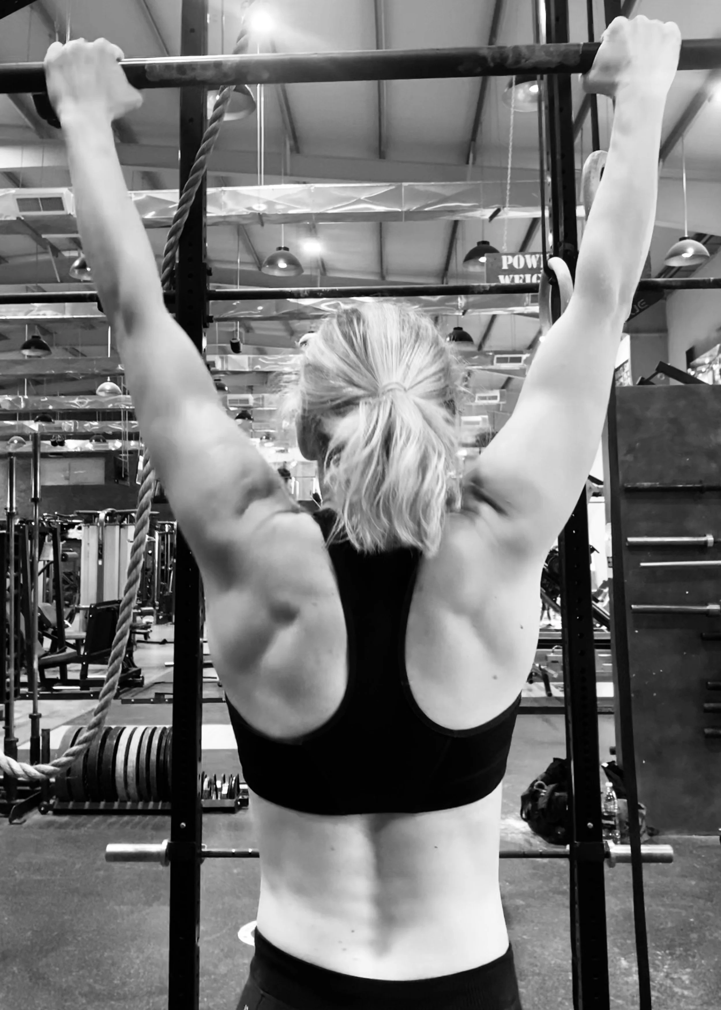 A woman with blonde hair in a ponytail is doing a pull-up exercise in a gym, hanging from a pull-up bar with both hands.