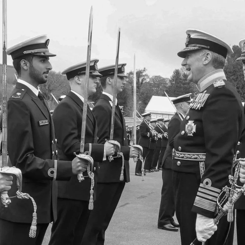 Military officers in uniform standing in formation during a formal event, with one officer smiling and facing others, outdoors with trees and tents in the background.