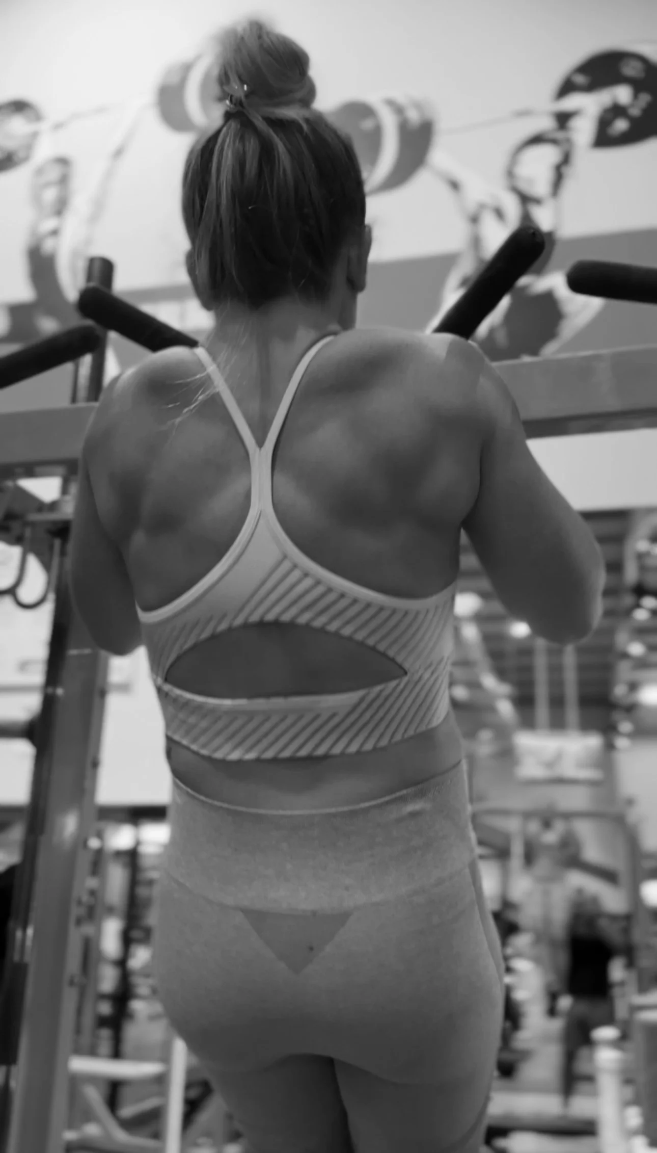 Back view of a woman in workout attire exercising in a gym, with weightlifting equipment and a mural in the background.