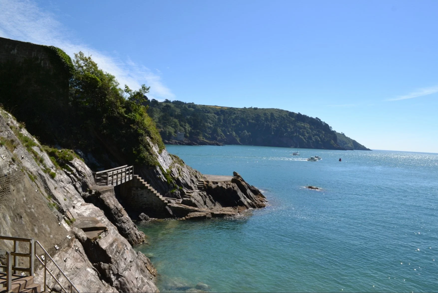 Coastal scene with rocky shoreline, stairs leading to water, green hillside, and boats on the calm blue sea under a clear sky.