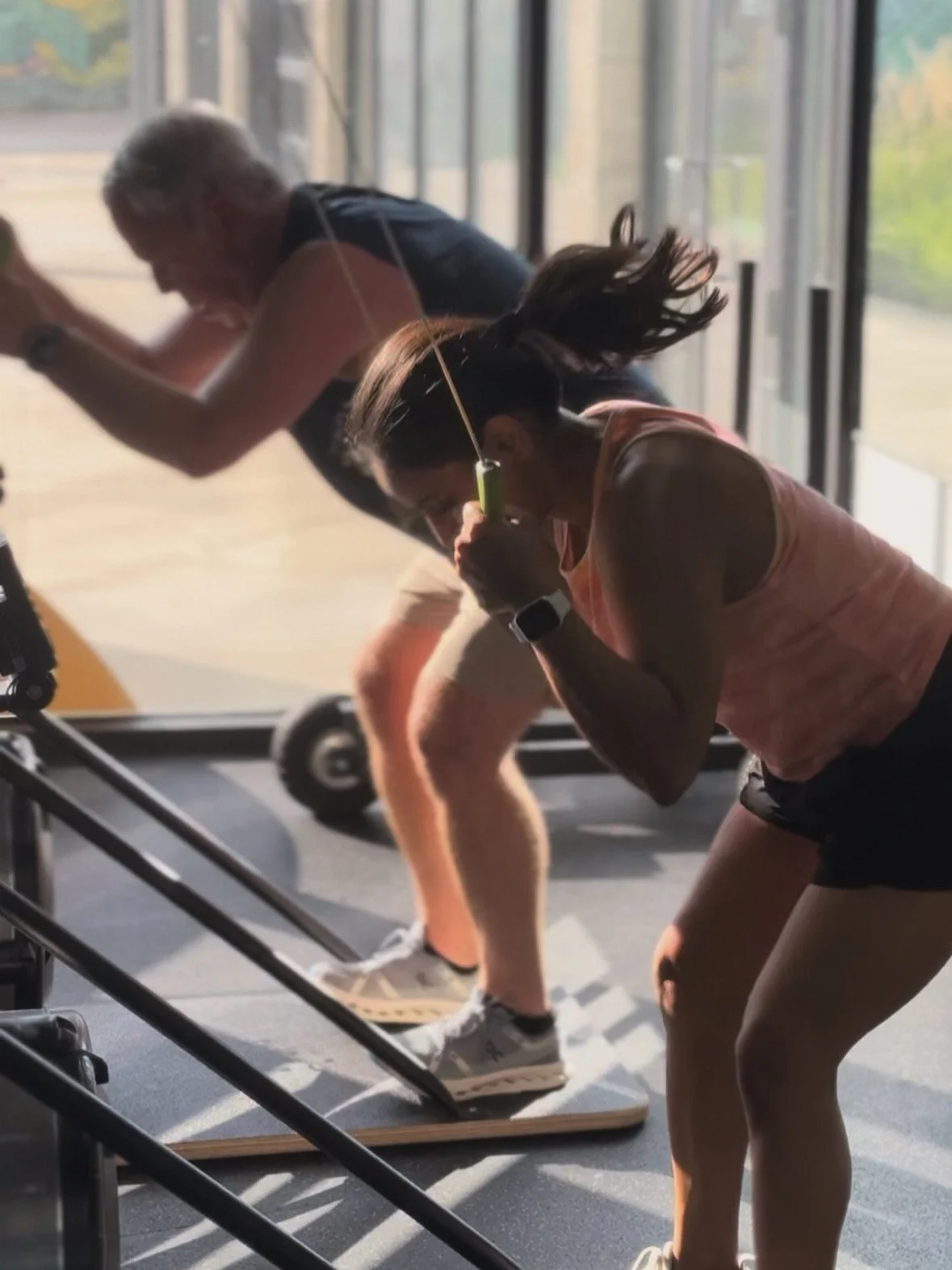 Two women exercise on elliptical machines in a gym, with sunlight coming through large windows.