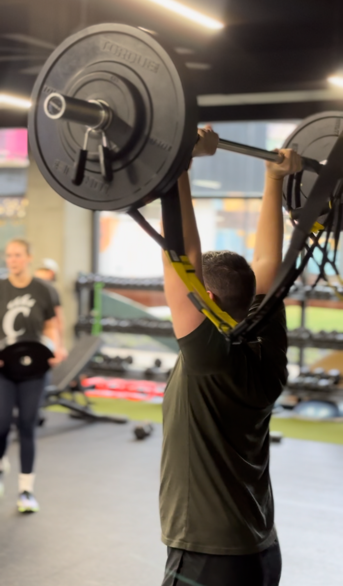 A person lifting a barbell with weights at a gym, with another person in the background.