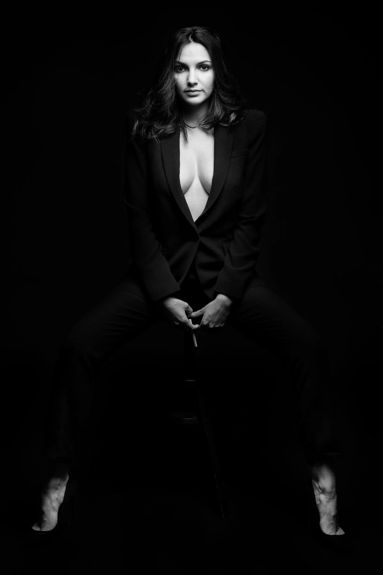 Une femme en costume noir, assise, avec un décolleté visible, positionnée contre un fond noir, en noir et blanc.