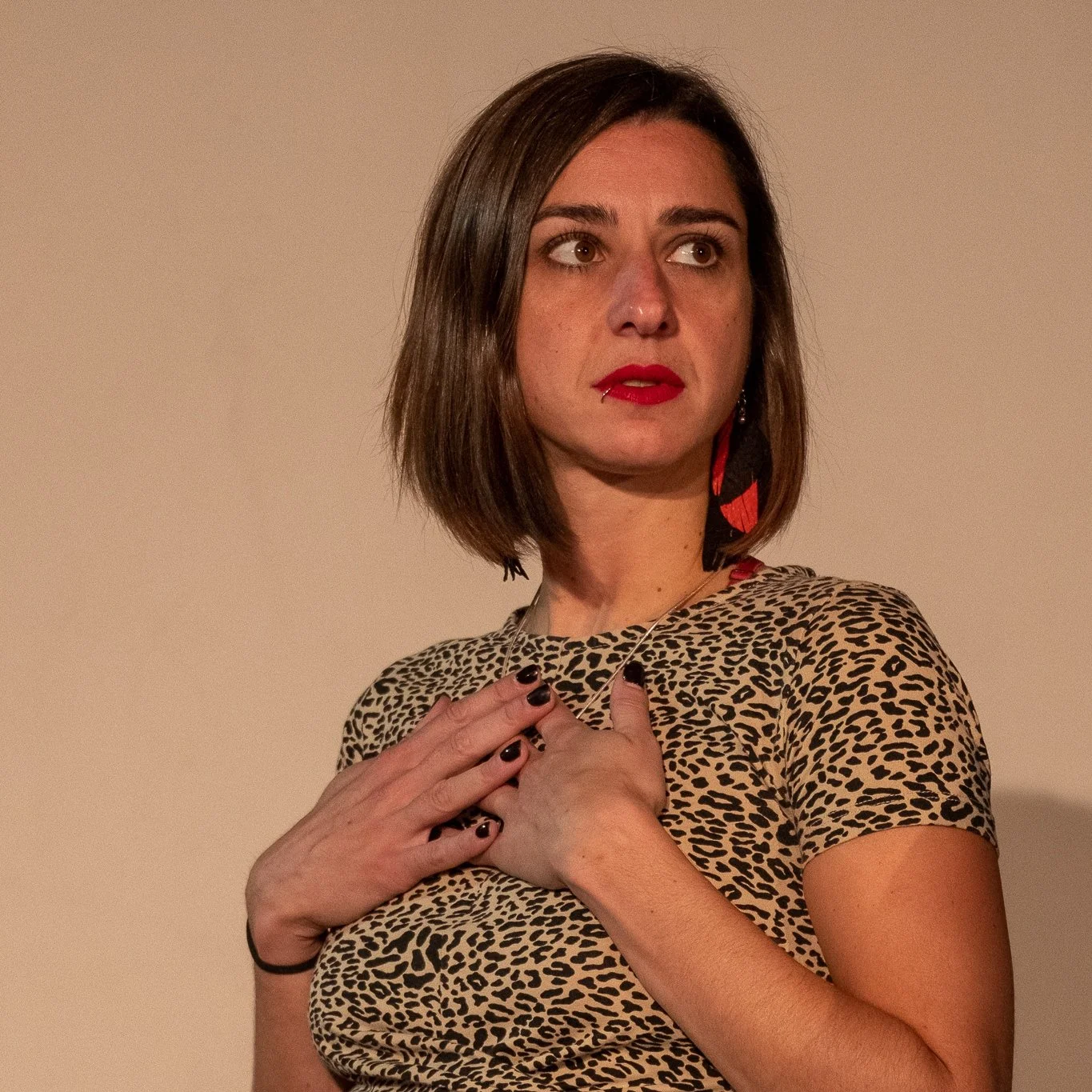 Billy Kissa with shoulder-length brown hair wearing a leopard print shirt, red lipstick, and large earrings. She has her hands crossed over her chest and appears to be looking to her right against a plain beige background.