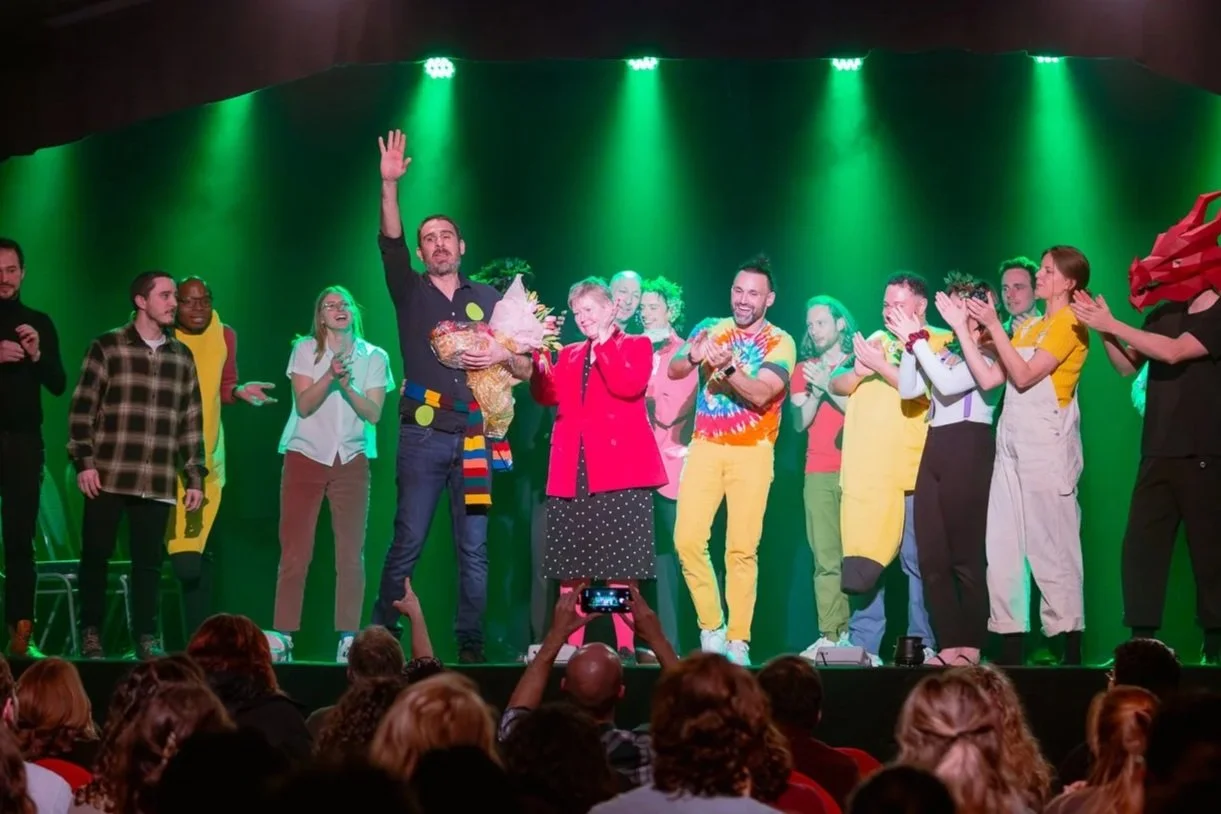 People on stage with green backdrop, some clapping and some holding flowers, and a person wearing a dragon mask on the right.