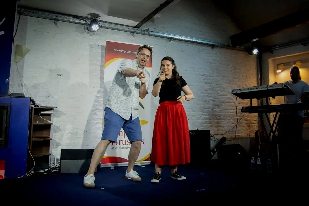 Two comedians, one male and one female, perform on stage at an improv comedy show. The male is wearing a white shirt and blue shorts, and the female is wearing a black top and red skirt. They are smiling and pointing towards the camera. A keyboard player is visible in the background to the right, and stage lights illuminate the scene.