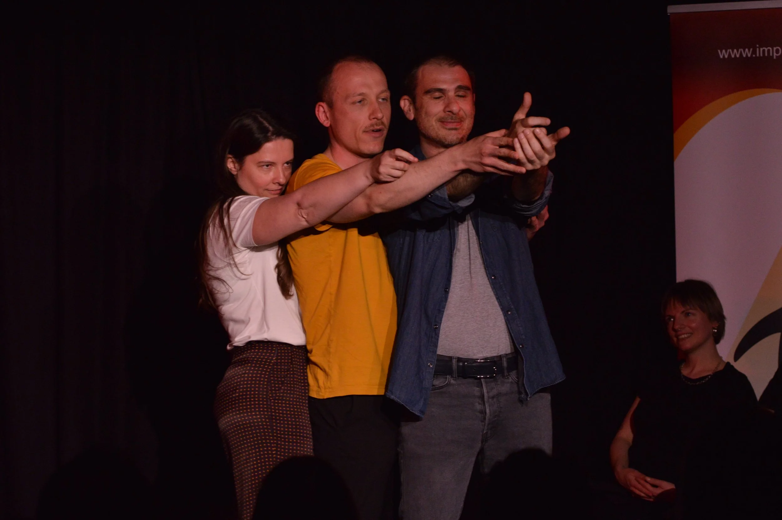 Three people standing closely, with their hands extended forward, appear to be participating in a shared activity or gesture on stage, with a woman smiling on the right and a banner in the background.