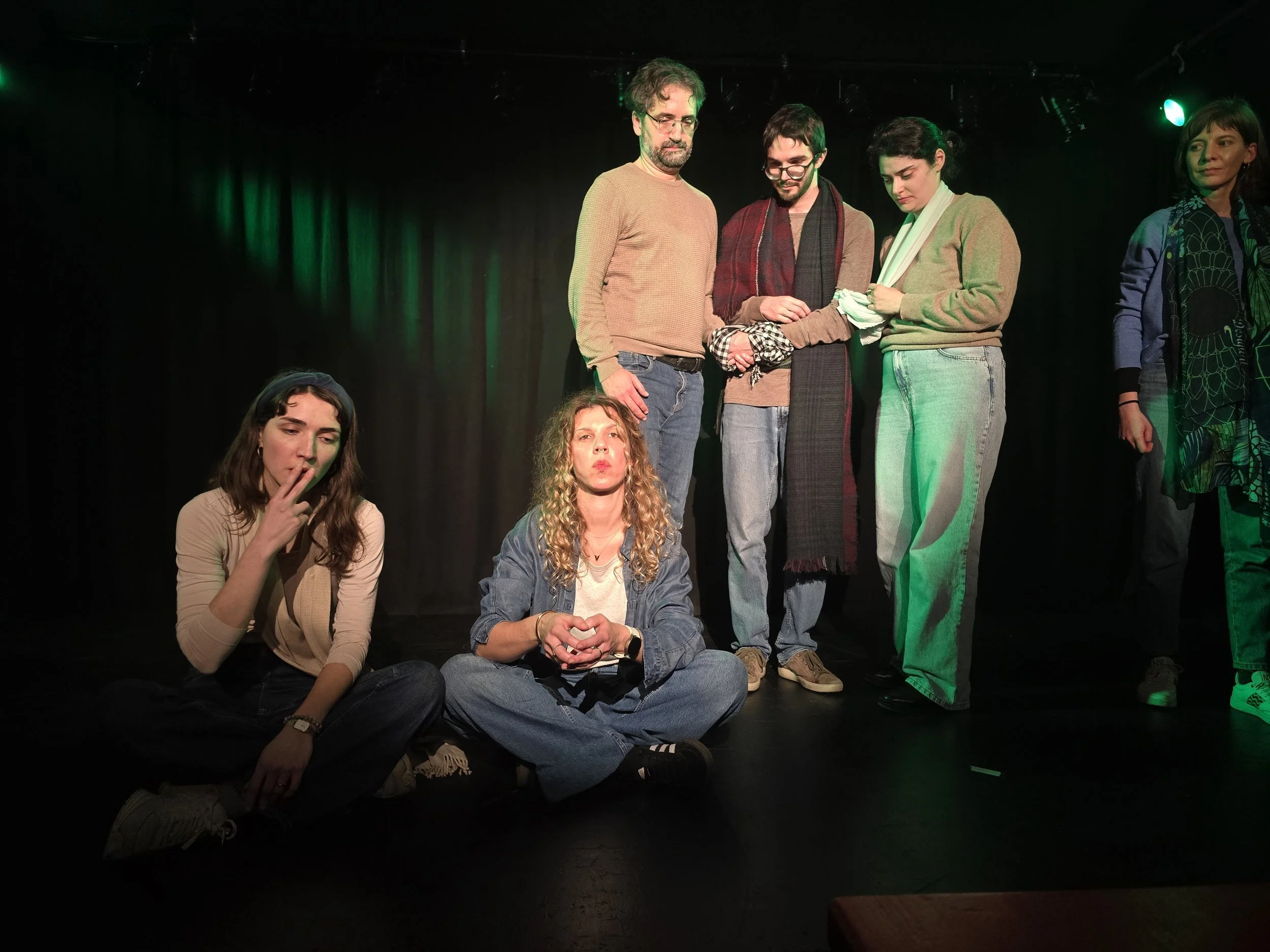 Group of five people on stage, three women sitting cross-legged at front, two men and one woman standing behind them, in a theater or performance space with black curtains and green stage lighting.