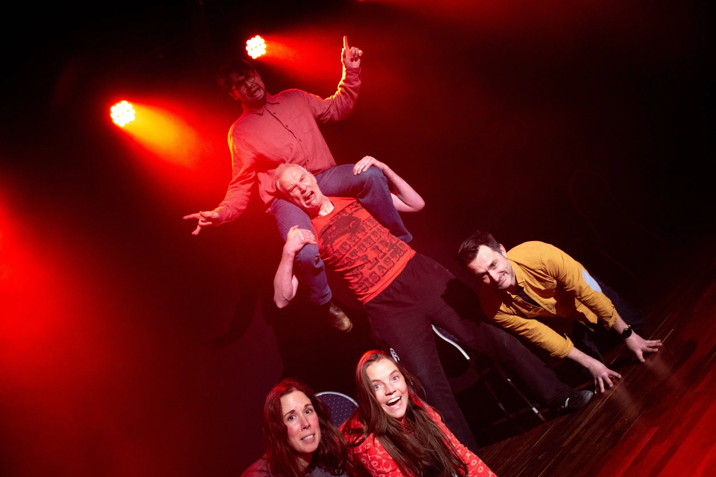 Impro Brussels students standing close together in a line, holding onto each other's shoulders or waist, performing a human pyramid or stacked formation under red stage lights on a dark stage.