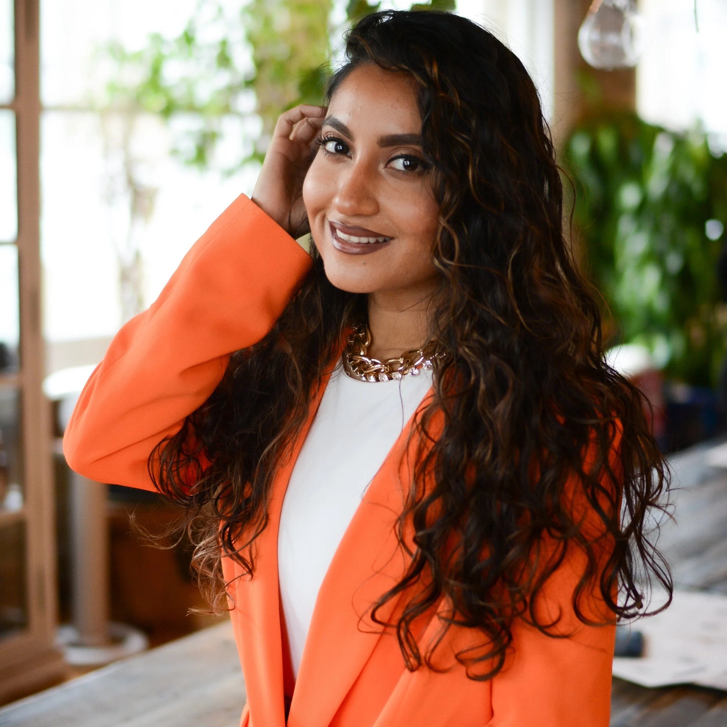 Woman with long curly hair wearing an orange blazer, smiling indoors.