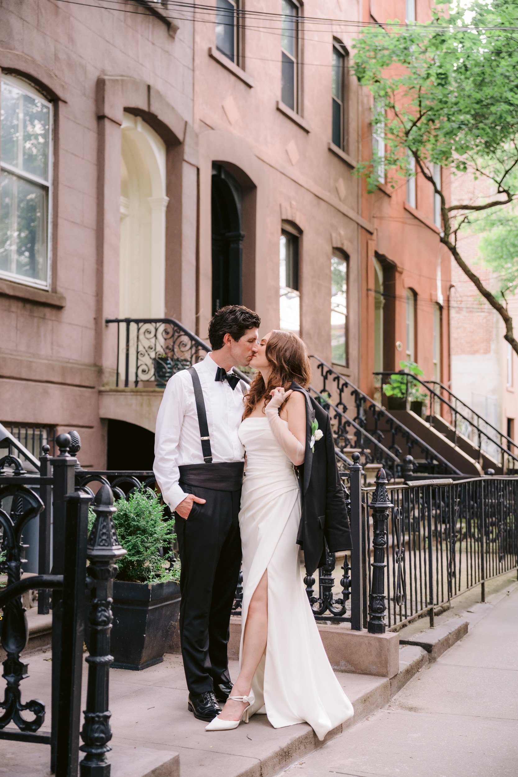 A couple kissing on a city sidewalk with brownstone buildings and green trees in the background, dressed in formal wedding attire.