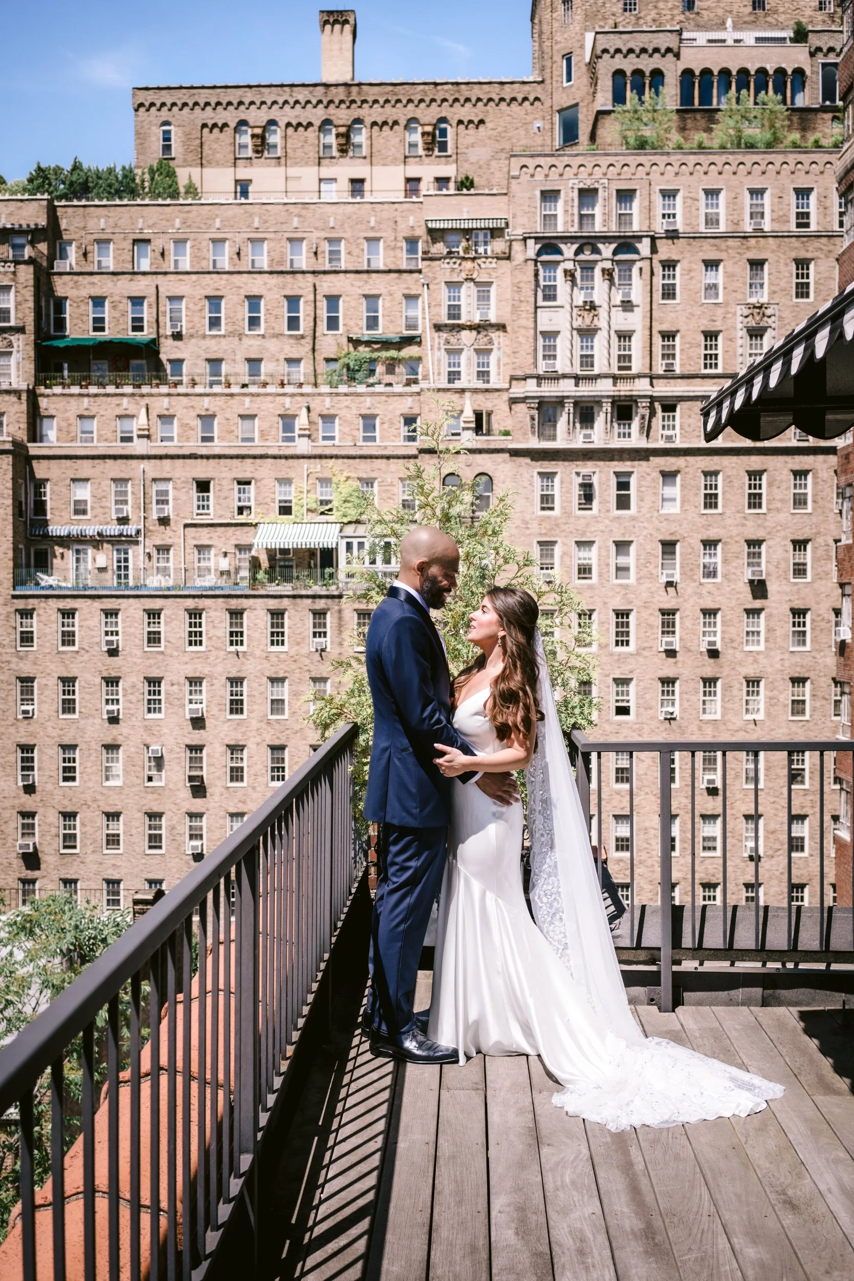 A bride and groom standing on a rooftop balcony, facing each other and holding hands, with an urban brick building in the background under a blue sky.