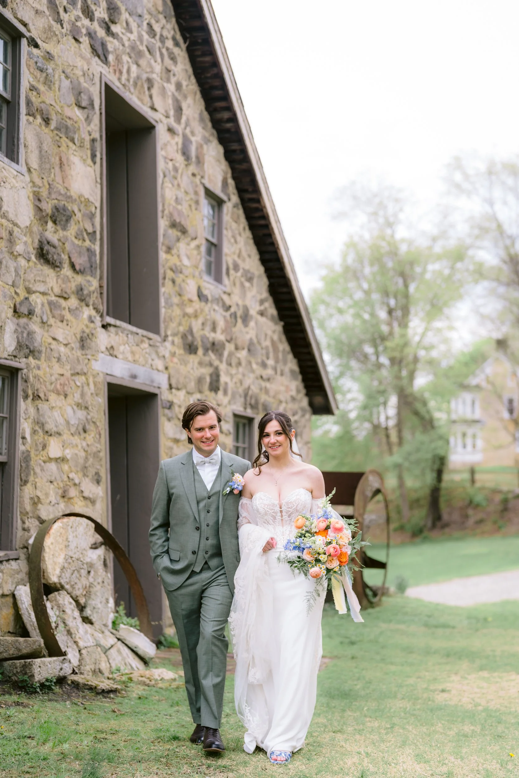 A bride and groom walking outdoors on their wedding day, with the bride holding a colorful bouquet of flowers. The groom is wearing a gray suit with a bow tie, and the bride is in a white lace wedding dress with long sleeves.