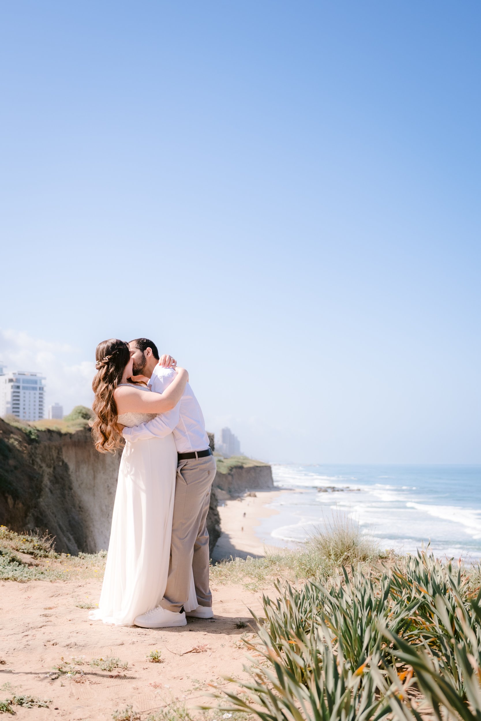A couple dressed in wedding attire kissing on a sandy beach with cliffs and the ocean in the background.