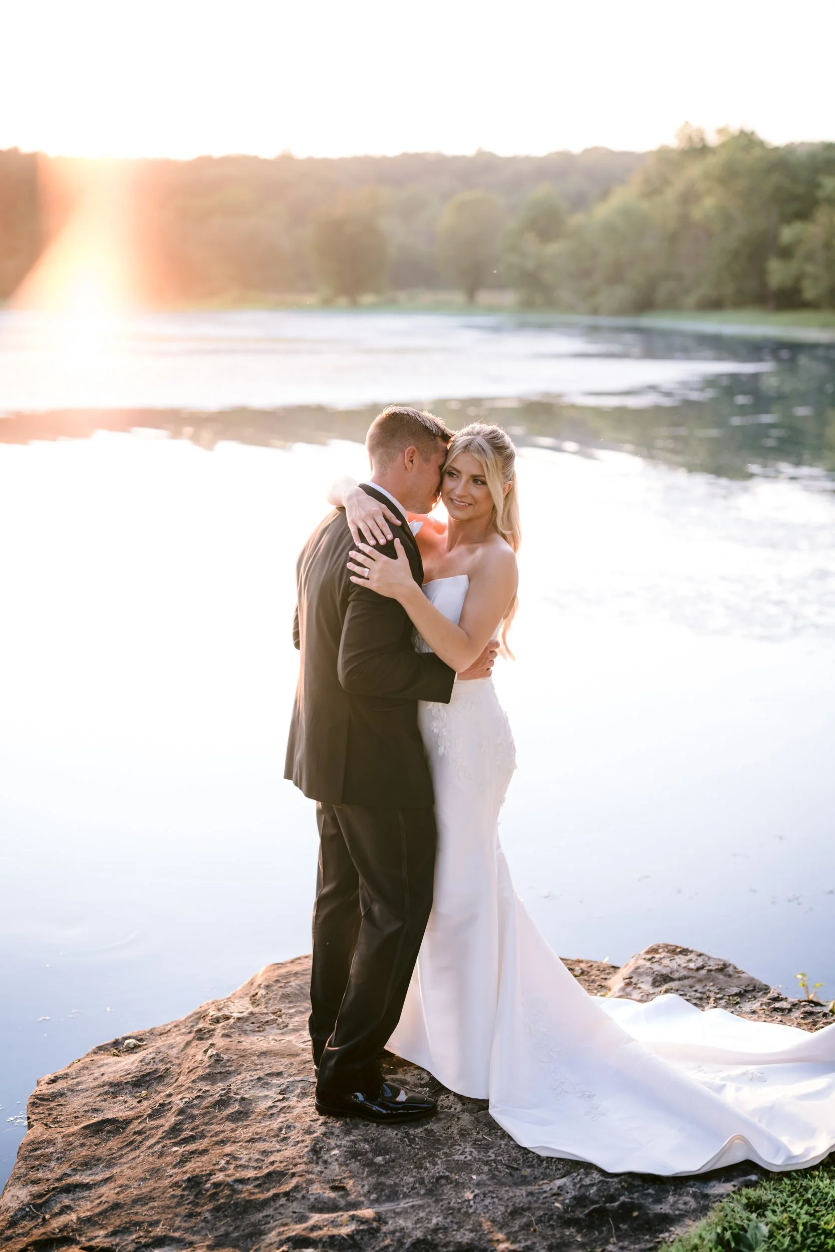 A bride and groom embrace by a river at sunset, the bride in a white wedding dress and the groom in a black tuxedo, standing on a rock with water and trees in the background.