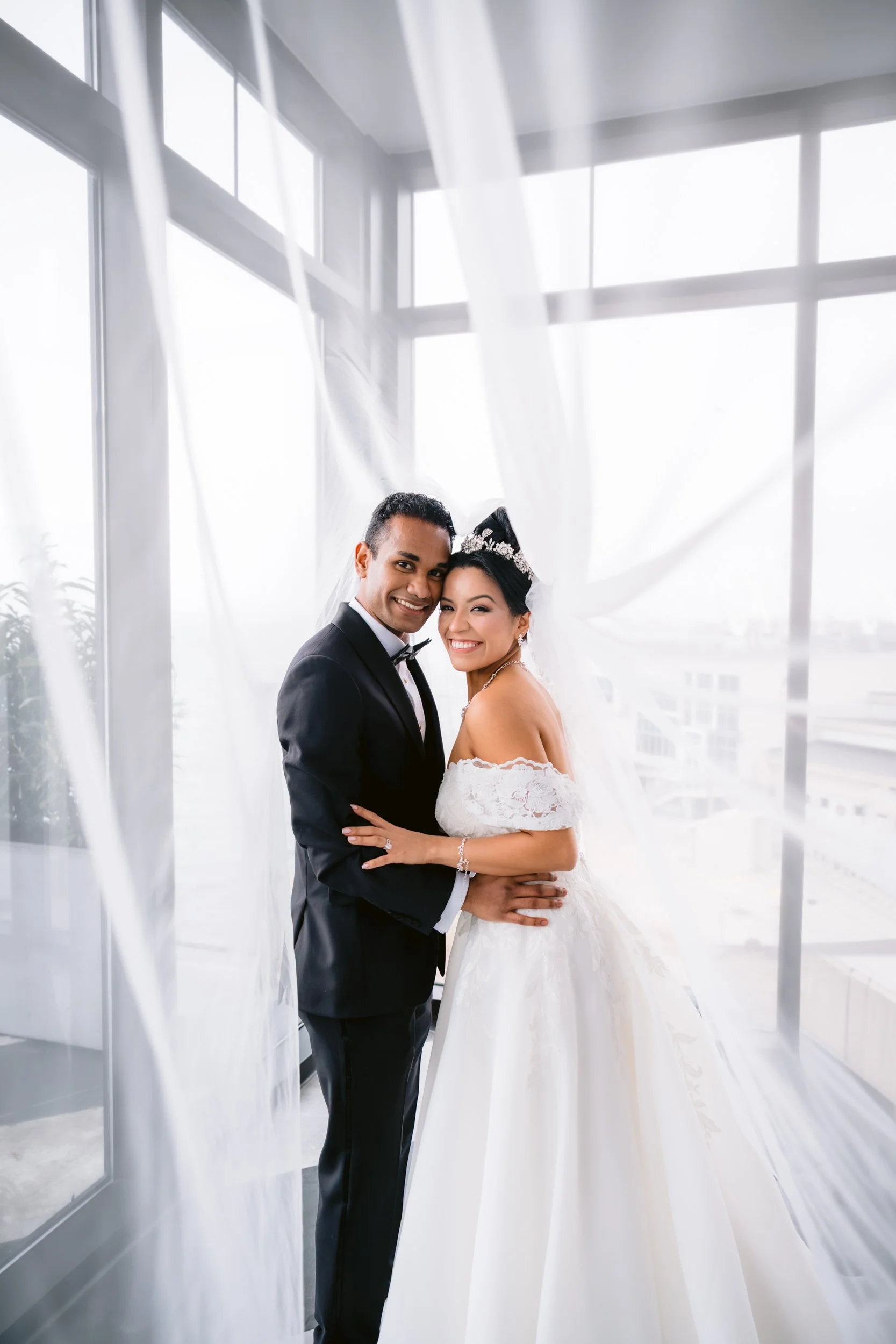 A happy couple dressed in wedding attire, the groom in a black tuxedo and the bride in a white wedding gown, standing close together in front of large windows with natural light.