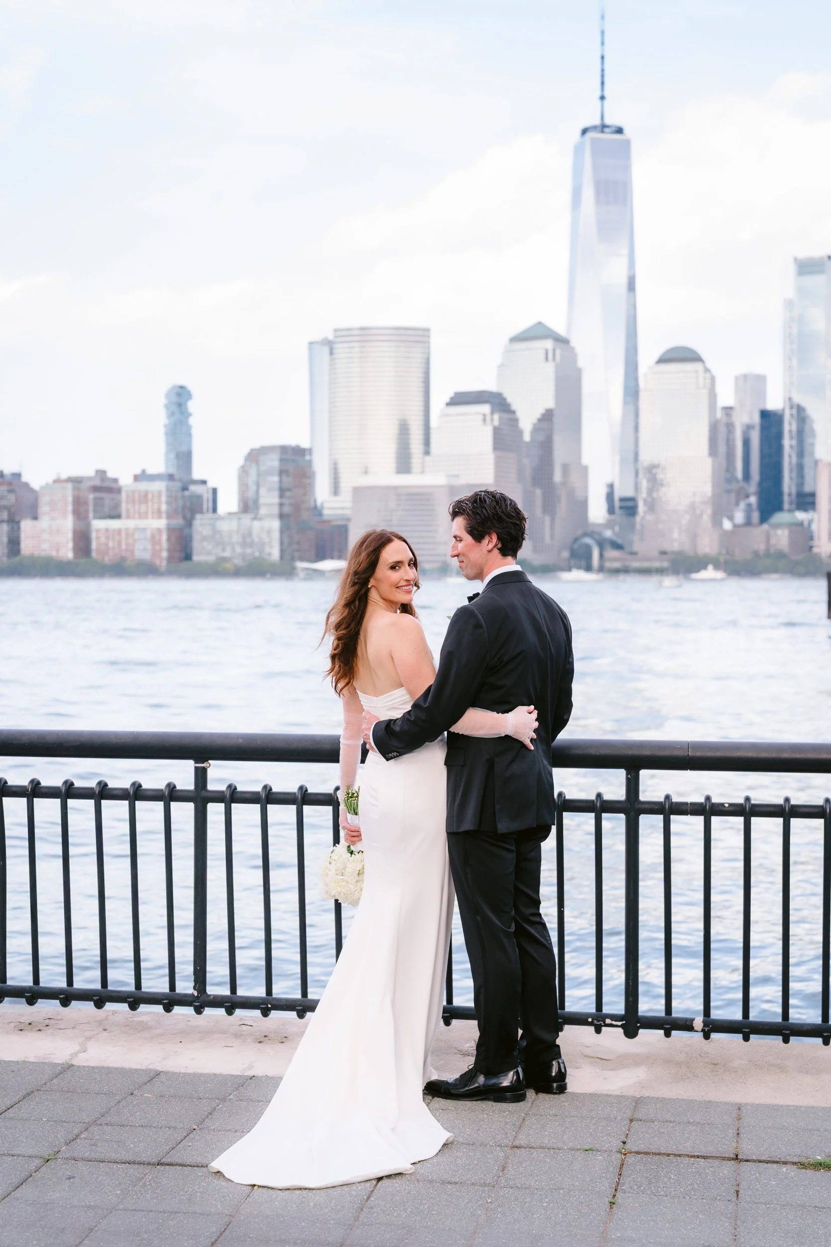 A bride and groom standing together by the water with a city skyline in the background, including the One World Trade Center in New York City.