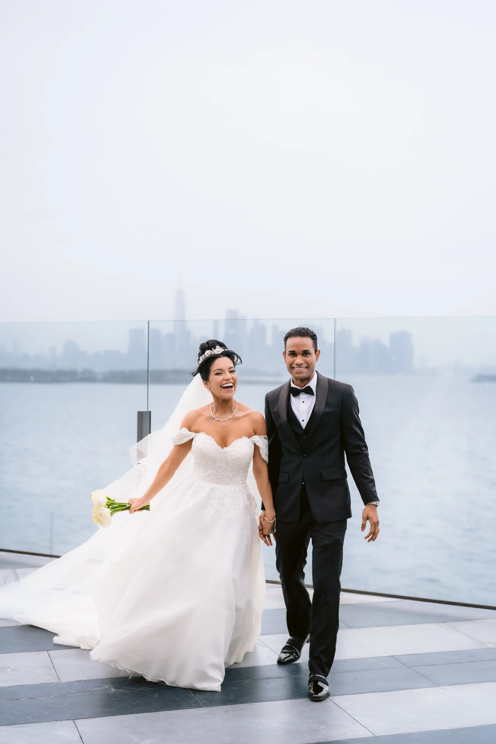 A smiling bride in a white wedding gown holding a bouquet of white roses, walking hand-in-hand with a smiling groom in a black tuxedo and bow tie on a rooftop with a city skyline in the background.