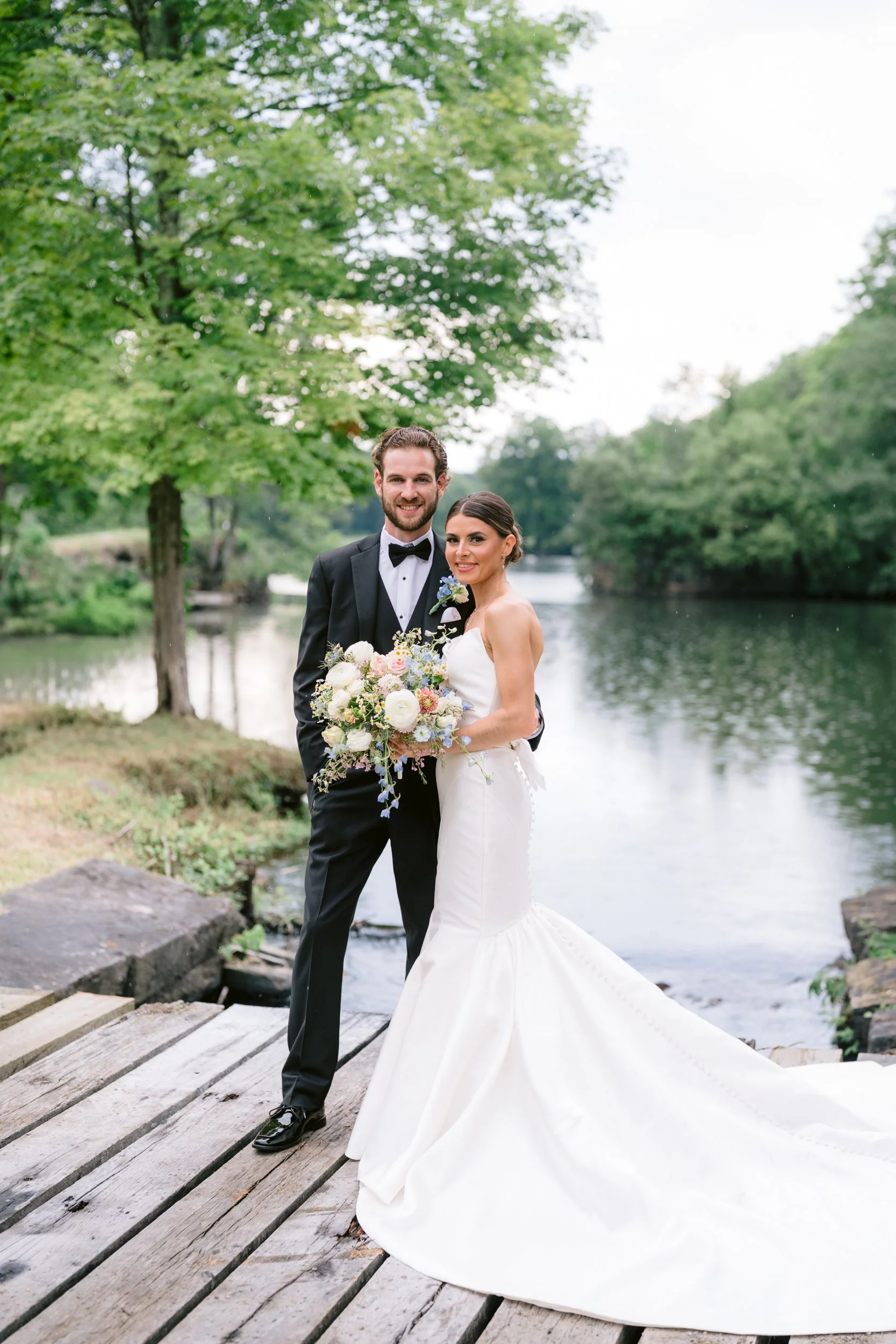 A newlywed couple stands on a wooden bridge by a river, surrounded by green trees. The groom wears a black tuxedo with a bow tie, and the bride wears a white wedding gown holding a bouquet of flowers. They are smiling and looking at the camera.