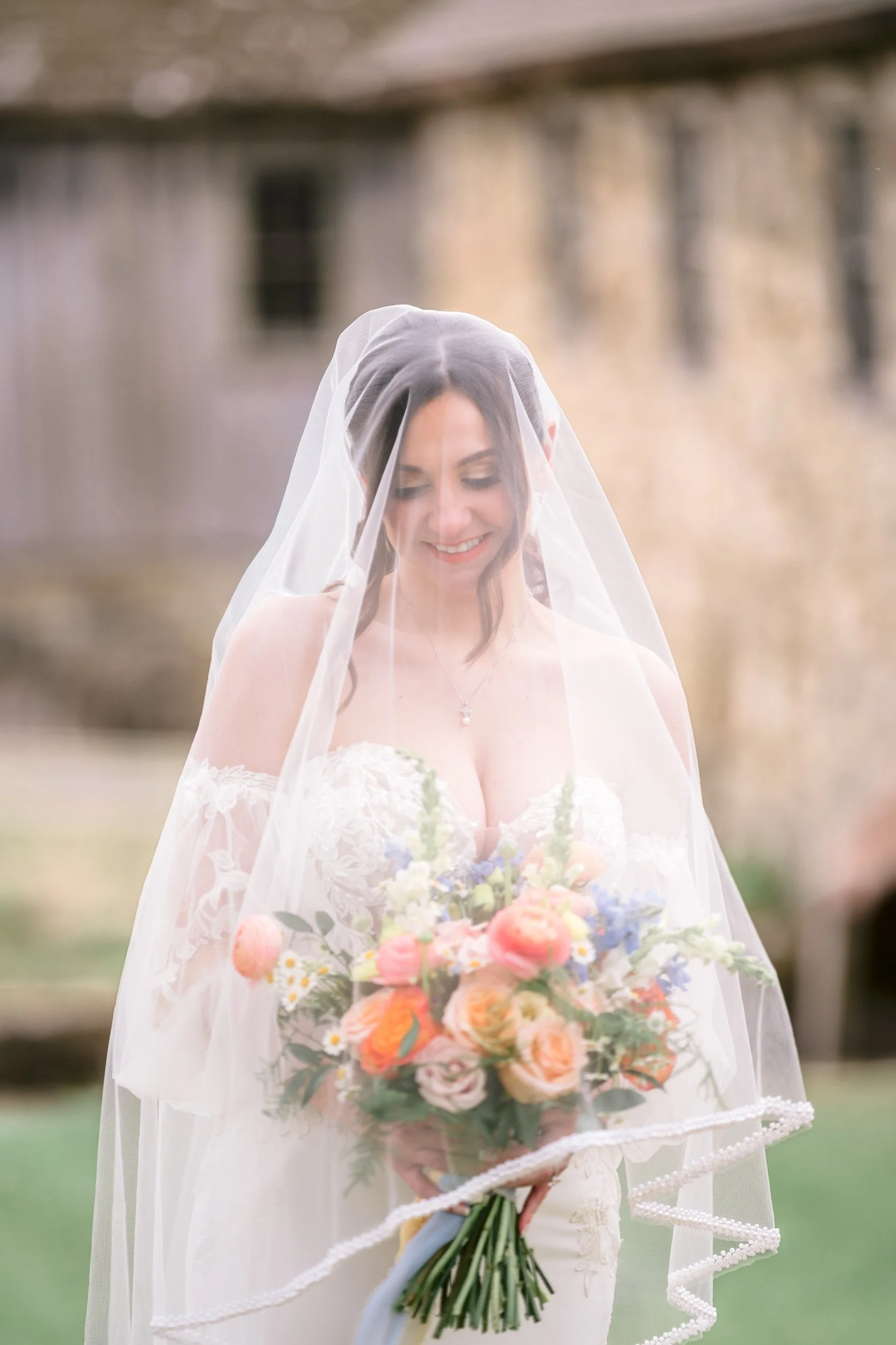 A bride in a white wedding dress and veil is holding a large bouquet of colorful flowers, standing outdoors with a blurred building in the background.