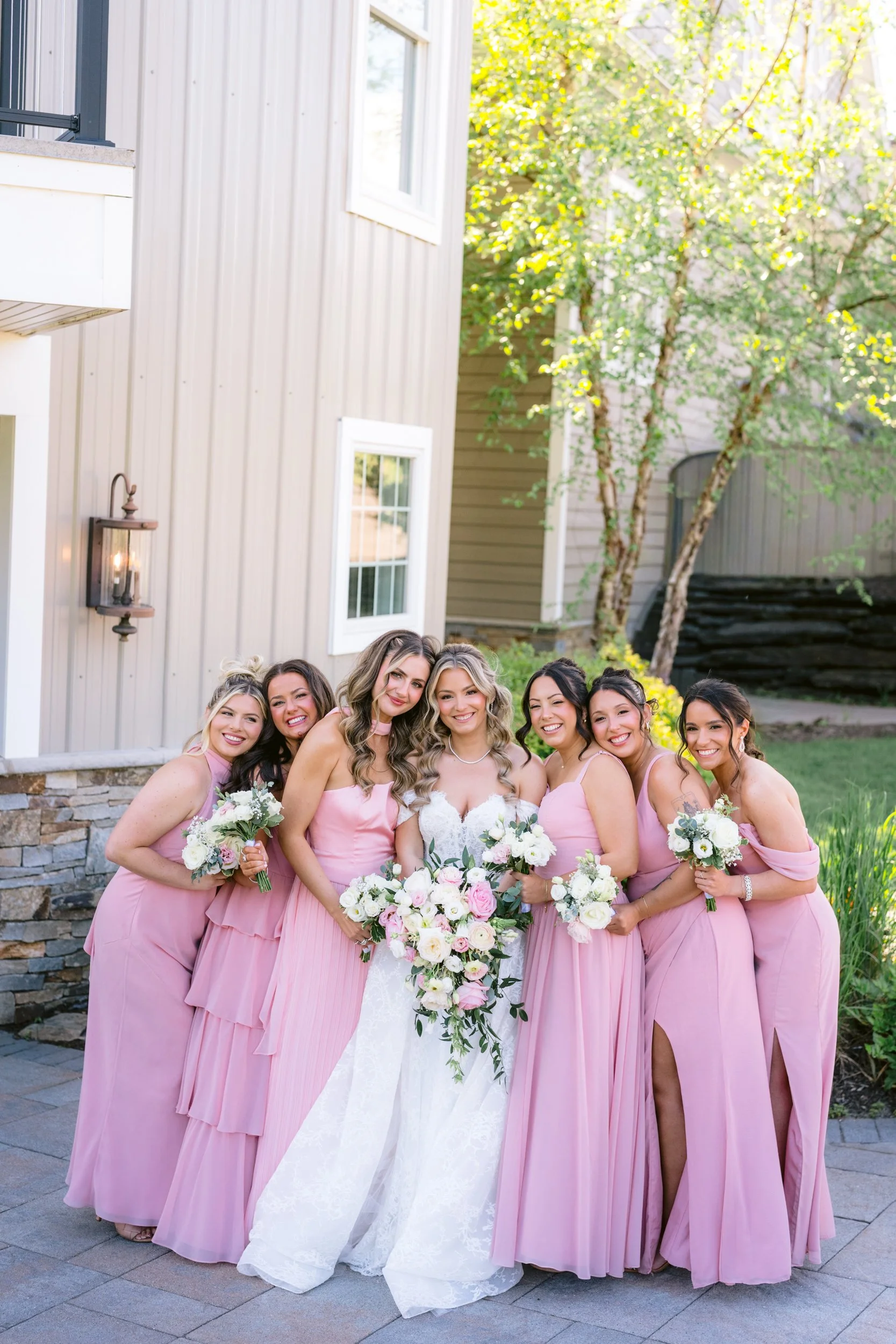 A group of women in pink dresses standing outdoors in front of a beige building with trees. Some women hold bouquets of white and pink flowers. They are smiling and looking at the camera.