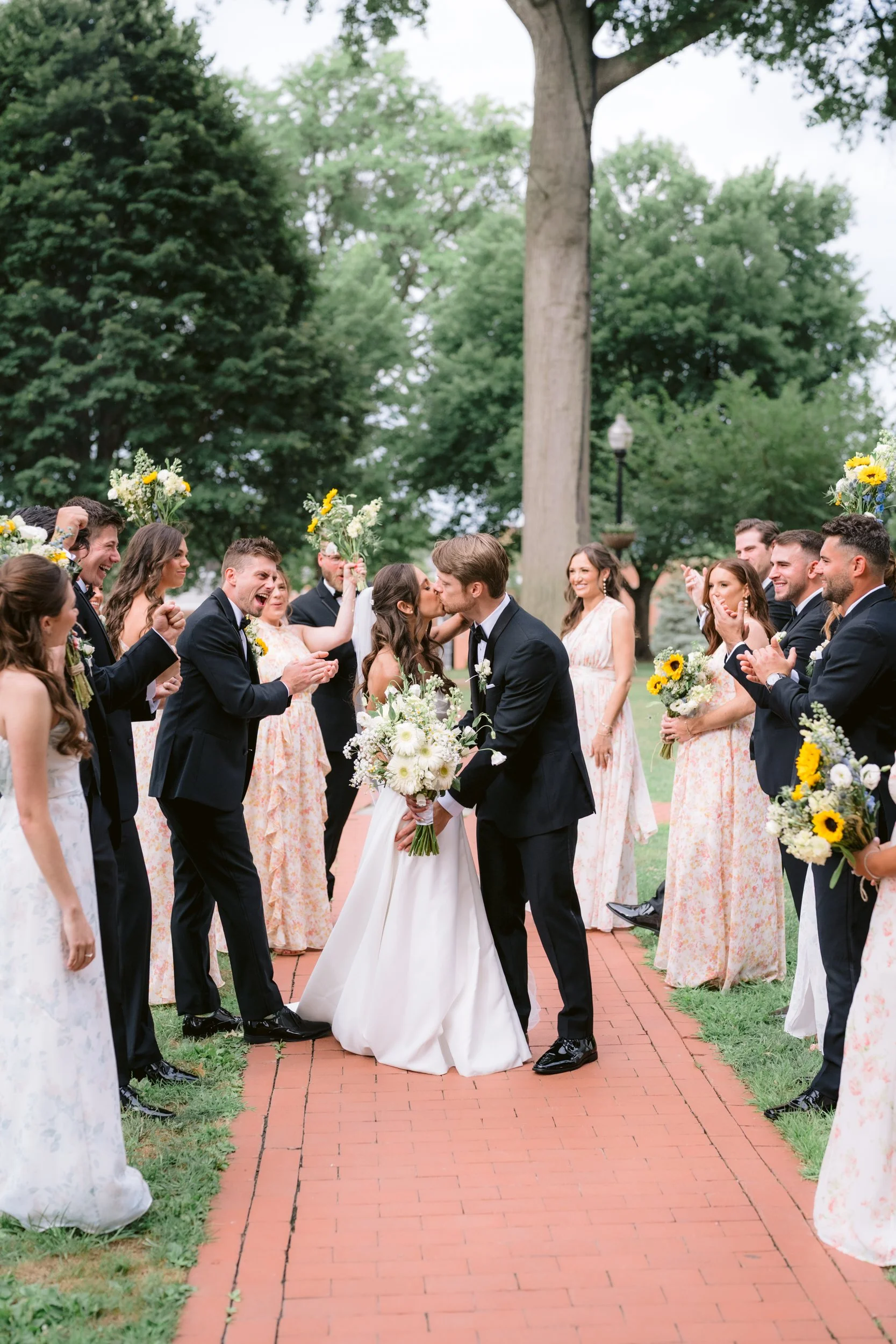 A wedding ceremony outdoors with a bride and groom kissing surrounded by friends and family, holding flowers, on a brick pathway with trees in the background.