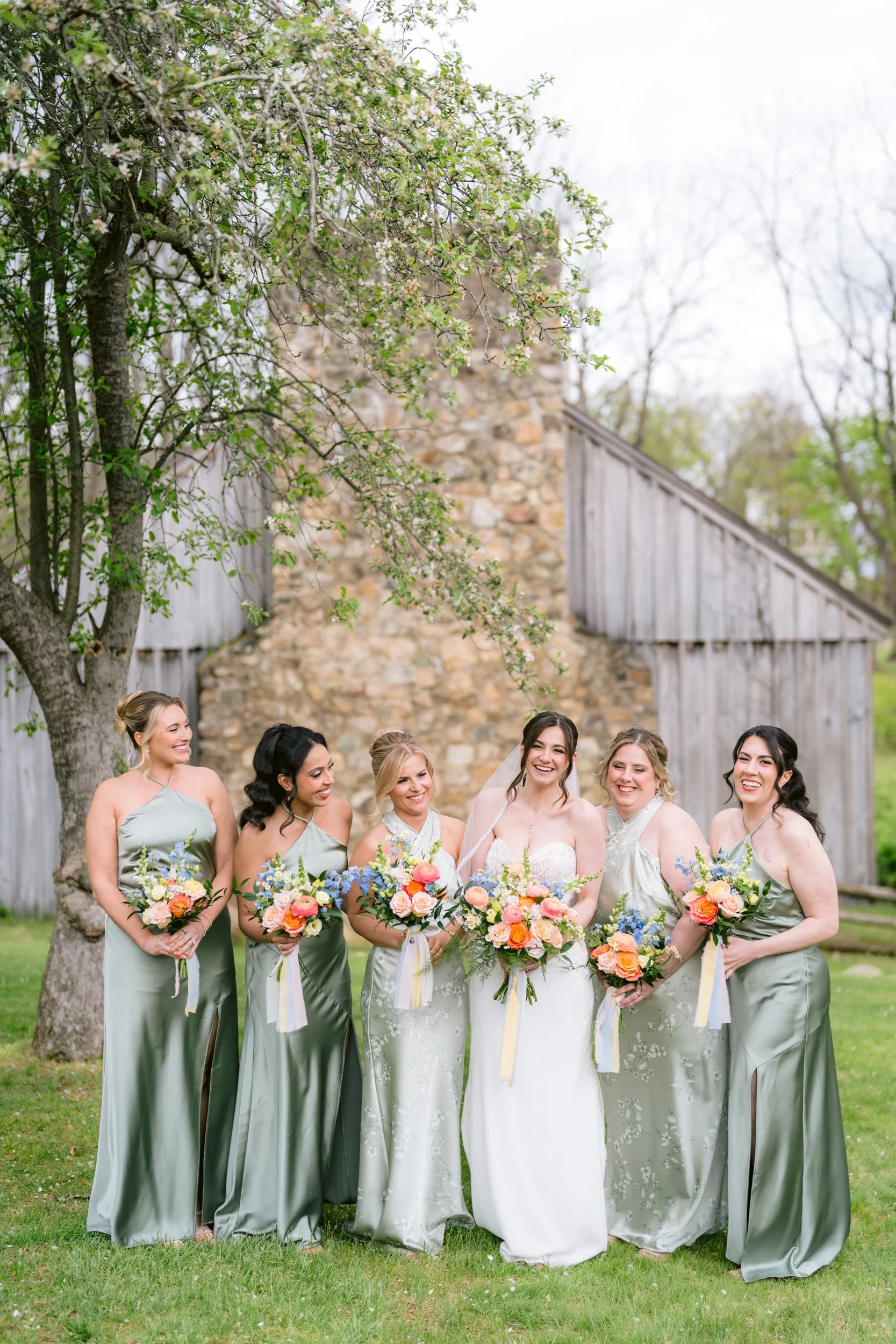 A group of bridal party members, including the bride and bridesmaids, standing outdoors on a grassy area with trees and a stone building in the background, all holding colorful bouquets, during a wedding celebration.
