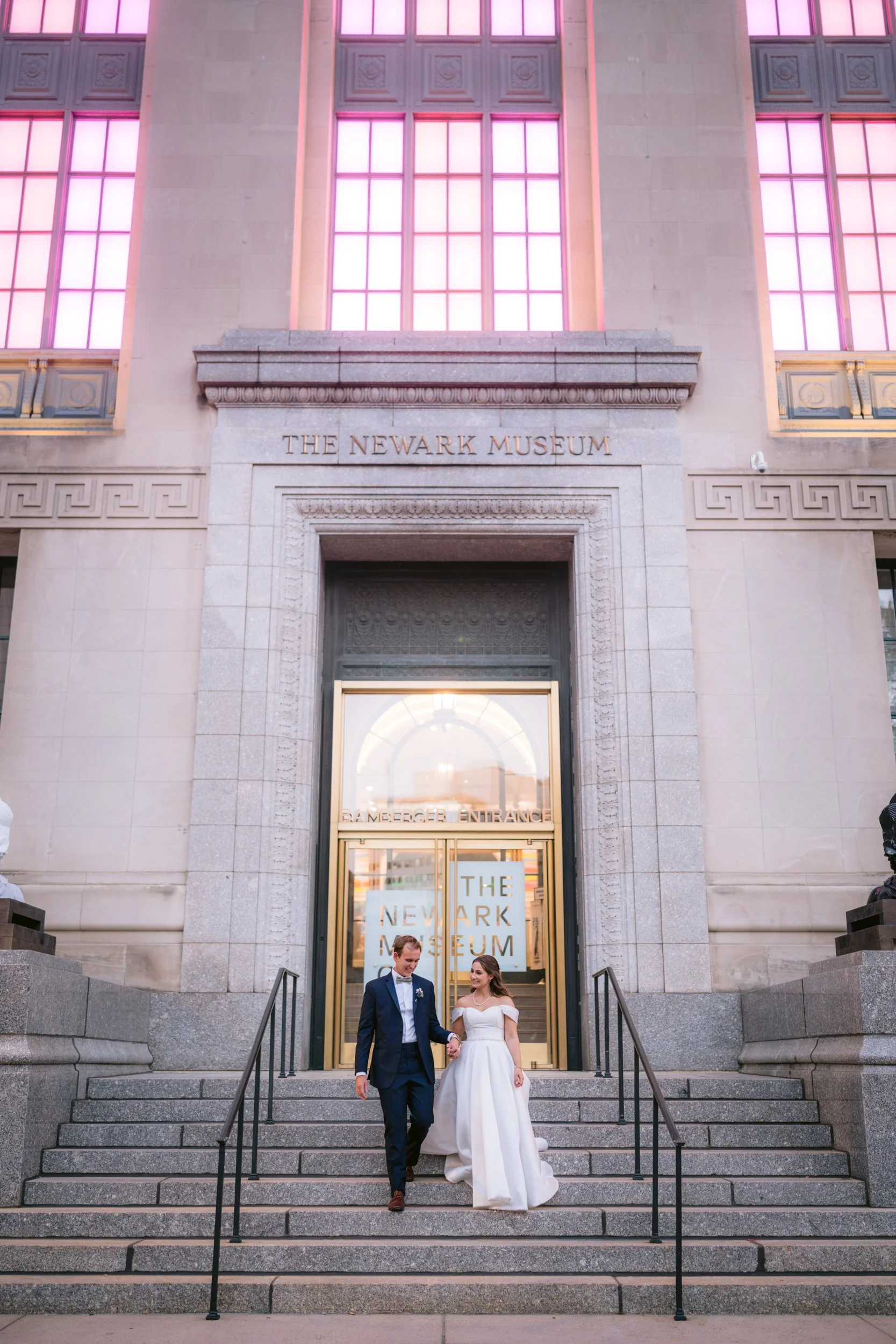 Couple in wedding attire walking down the steps outside the Newark Museum, with the museum's entrance behind them.