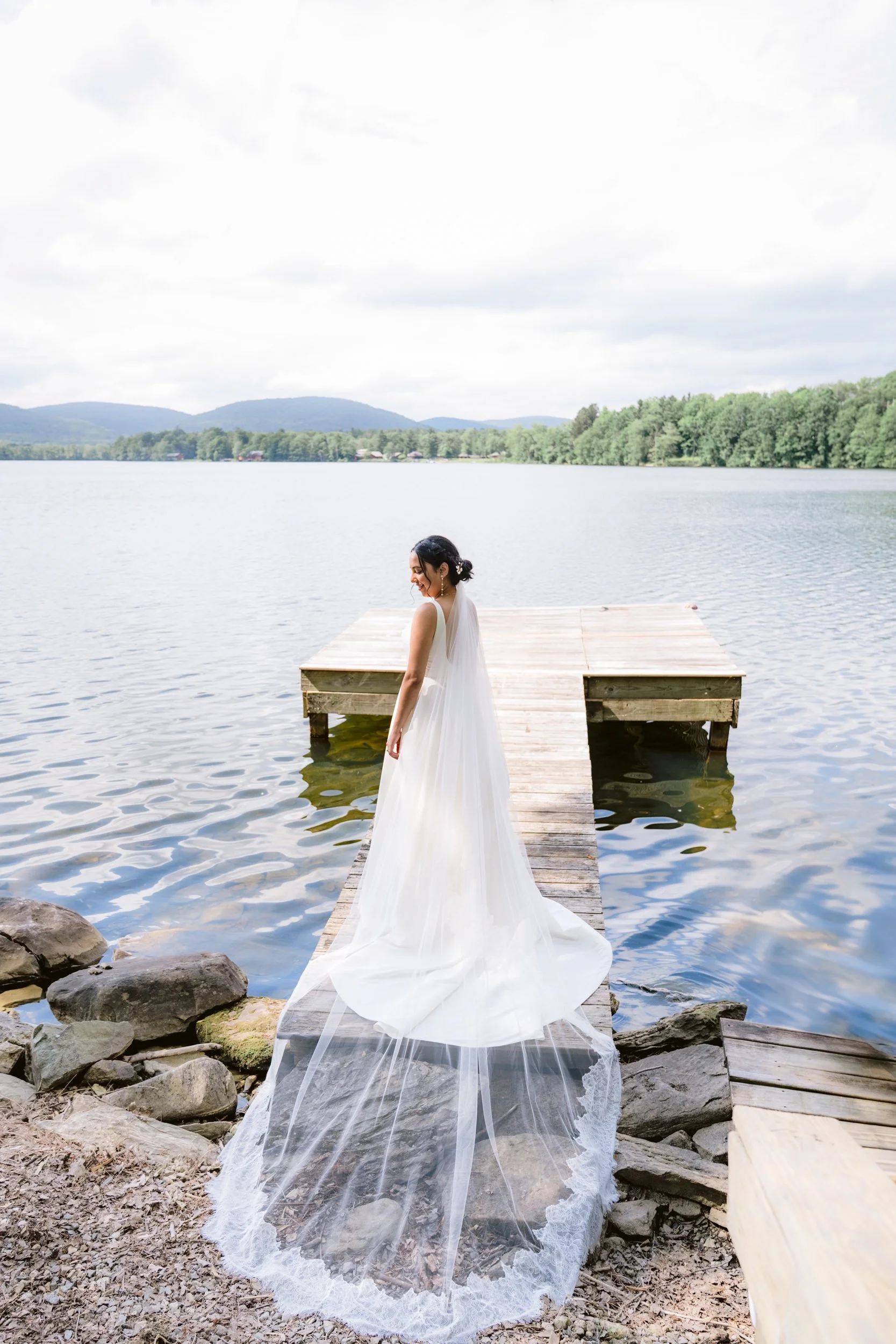 A woman in a white wedding dress standing on a wooden dock by a lake, with mountains and greenery in the background.