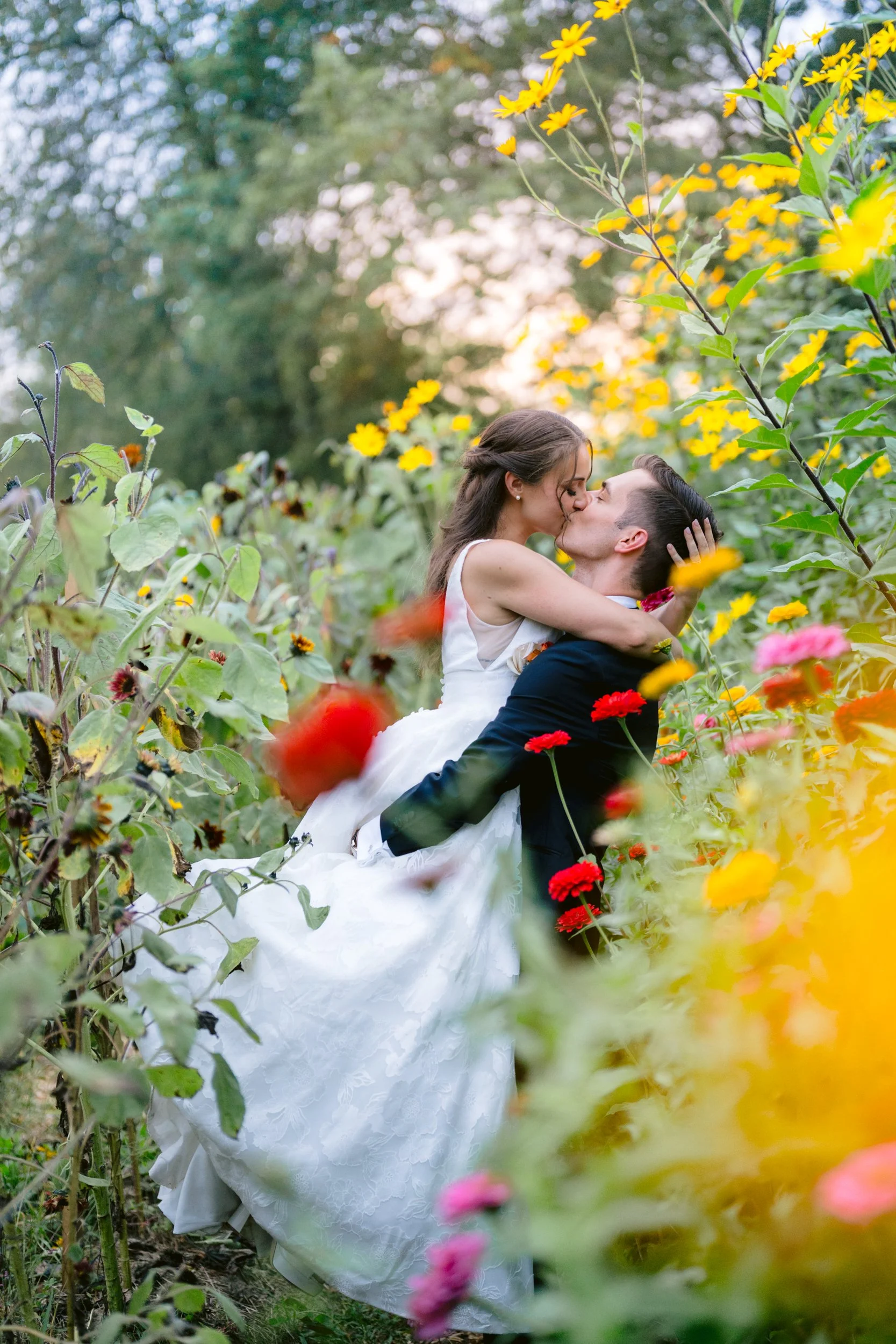A couple in wedding attire kissing in a garden with yellow, pink, and orange flowers surrounding them.