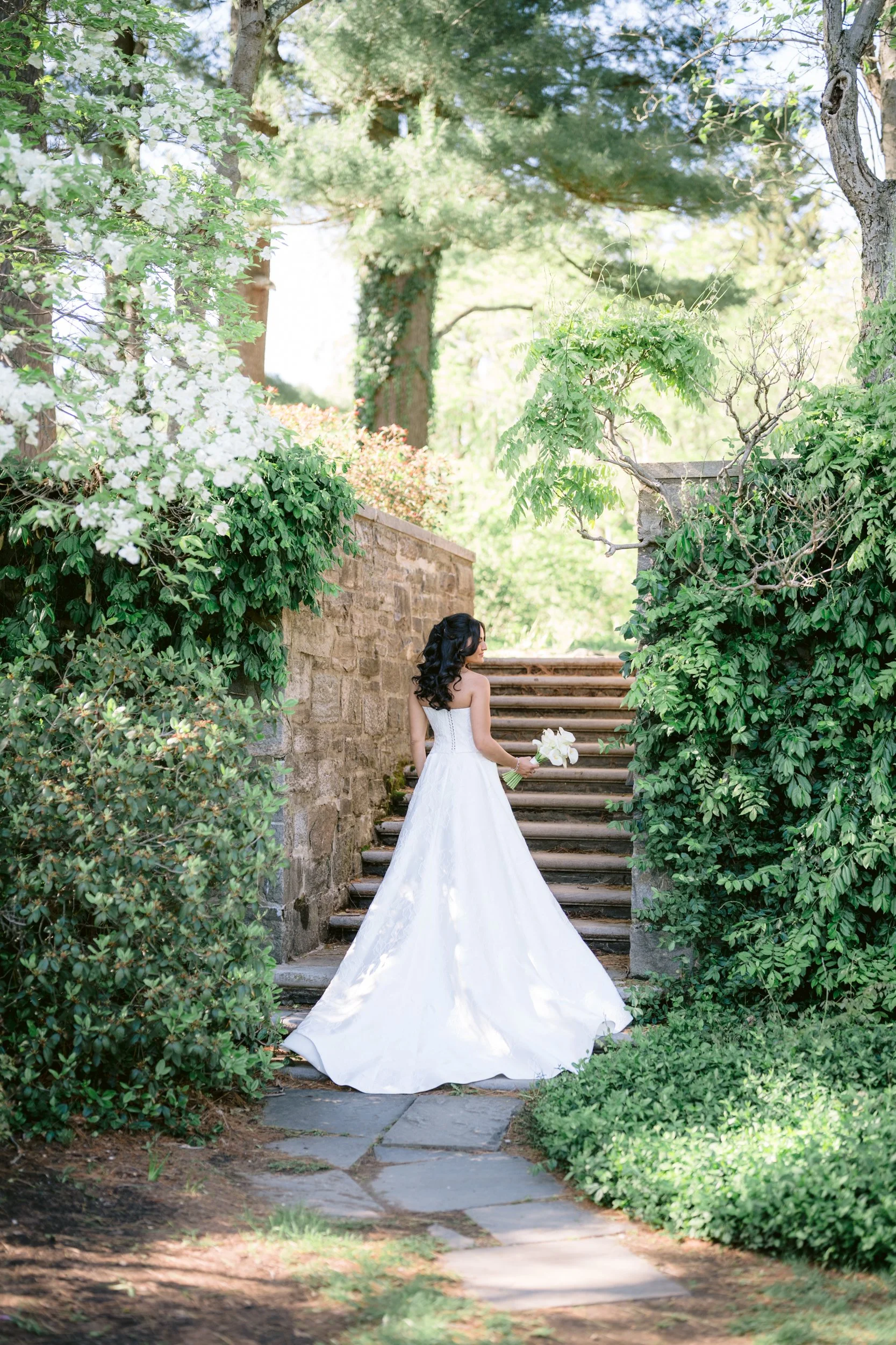 A bride in a white wedding gown holding a bouquet standing on stone steps surrounded by lush green shrubs and trees in a garden setting.
