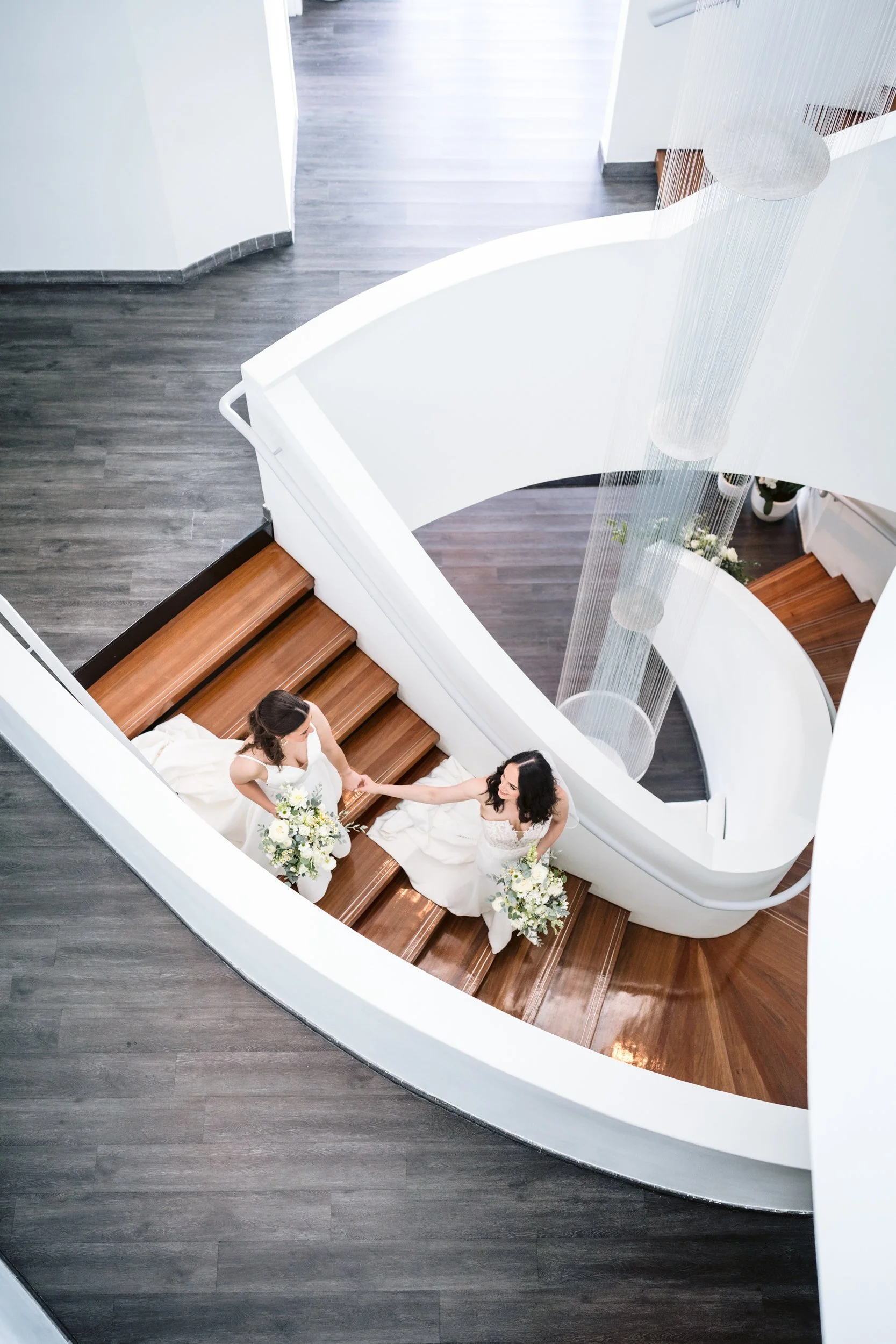 Two women in wedding dresses holding hands and bouquets, standing on a wooden staircase in a modern building with white walls and a curved design.