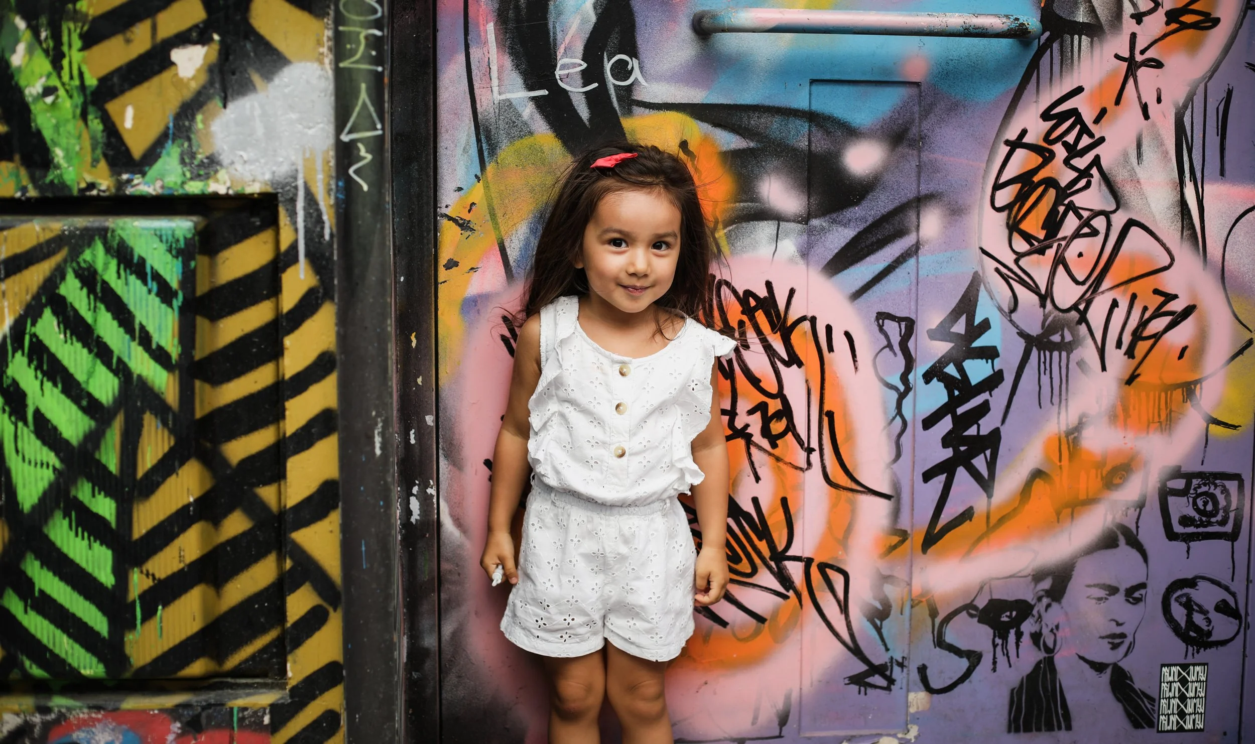 Jeune fille souriante en vêtements blancs avec un ruban rose dans les cheveux, se tenant devant un mur de graffiti coloré et artistique.