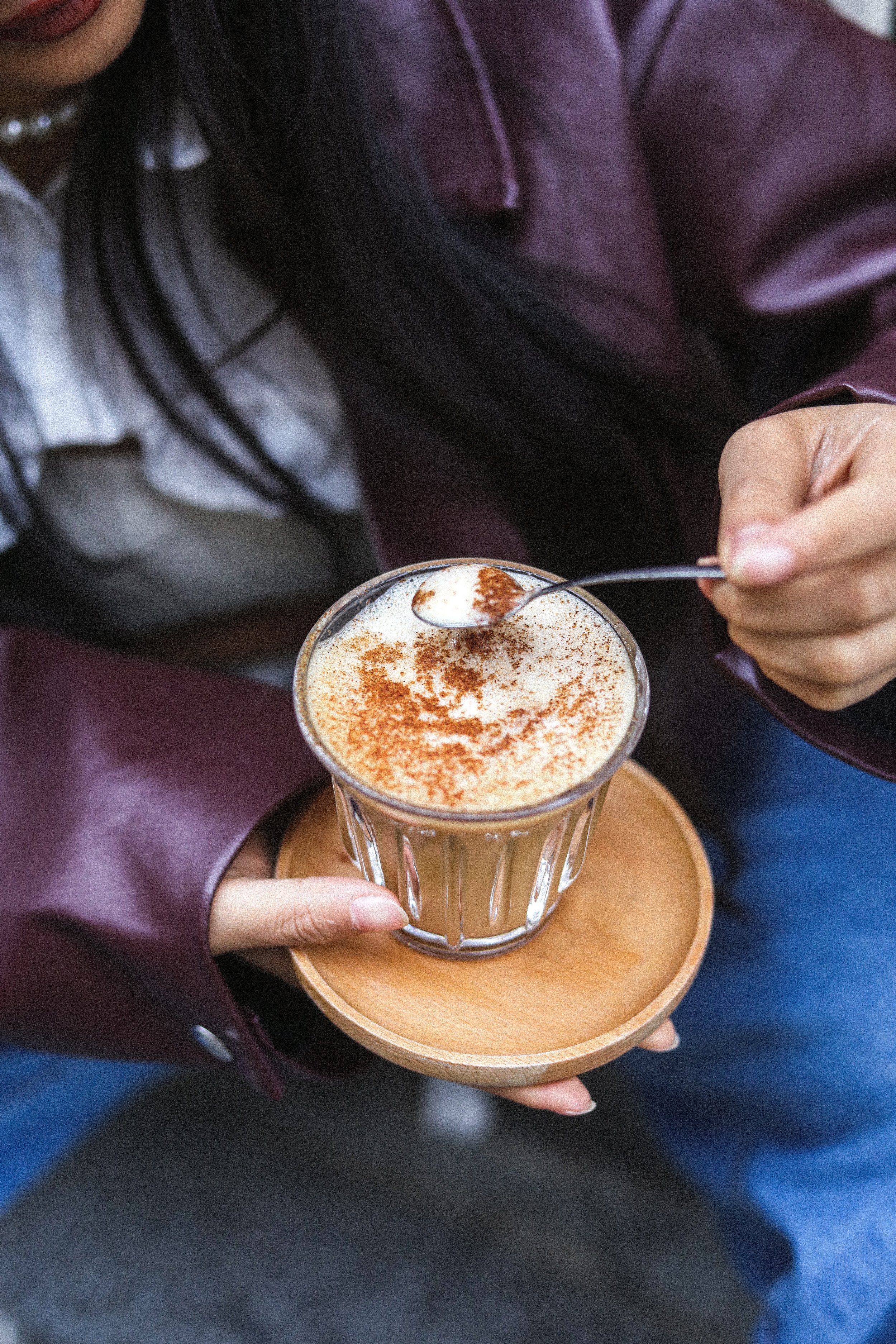 Une personne tient une petite tasse en verre contenant un café, garnie de cacao en poudre, sur une petite assiette en bois.