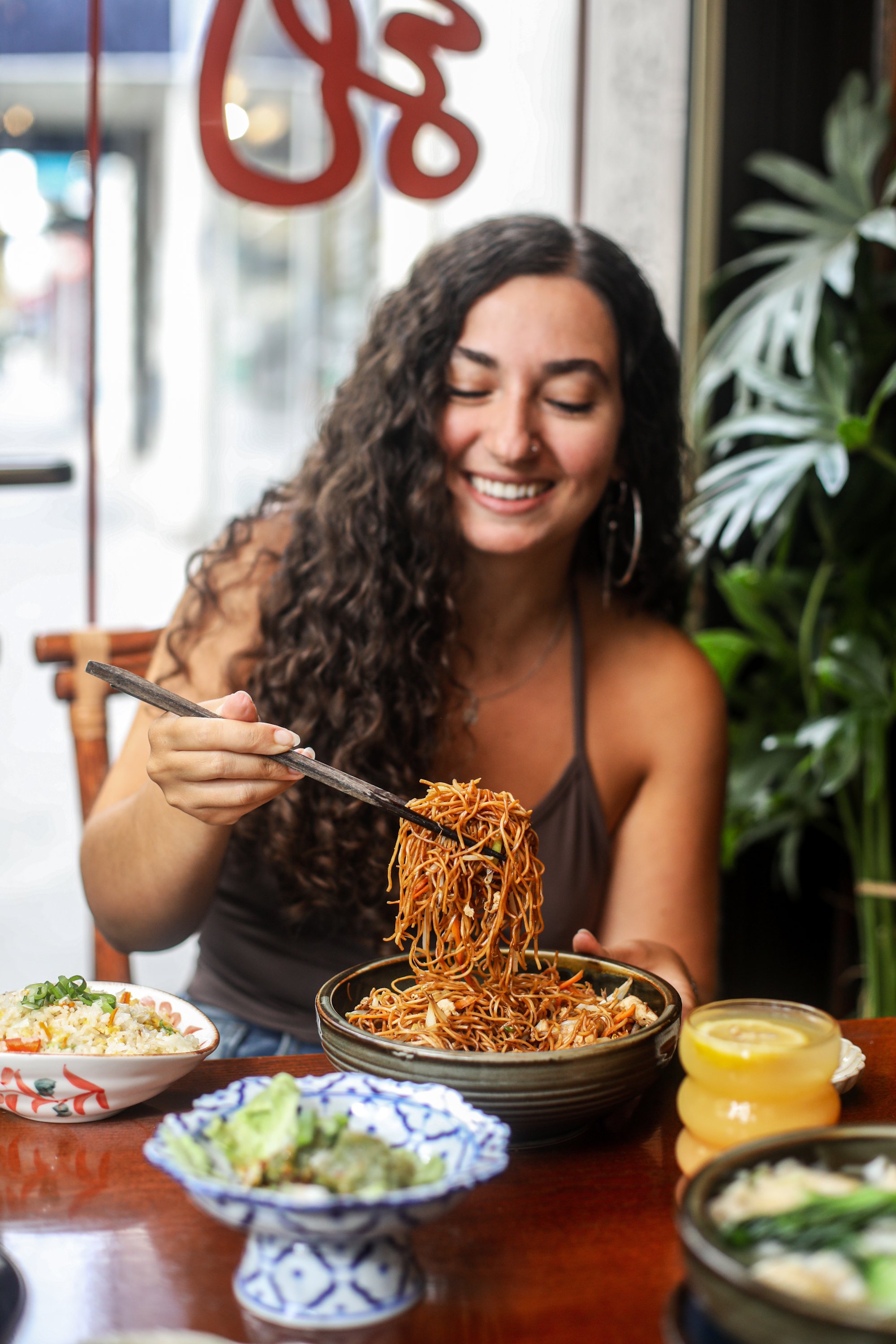 Femme souriante mangeant des nouilles dans un restaurant, avec des plats de cuisine asiatique sur la table, décor vertueux et lumineux.