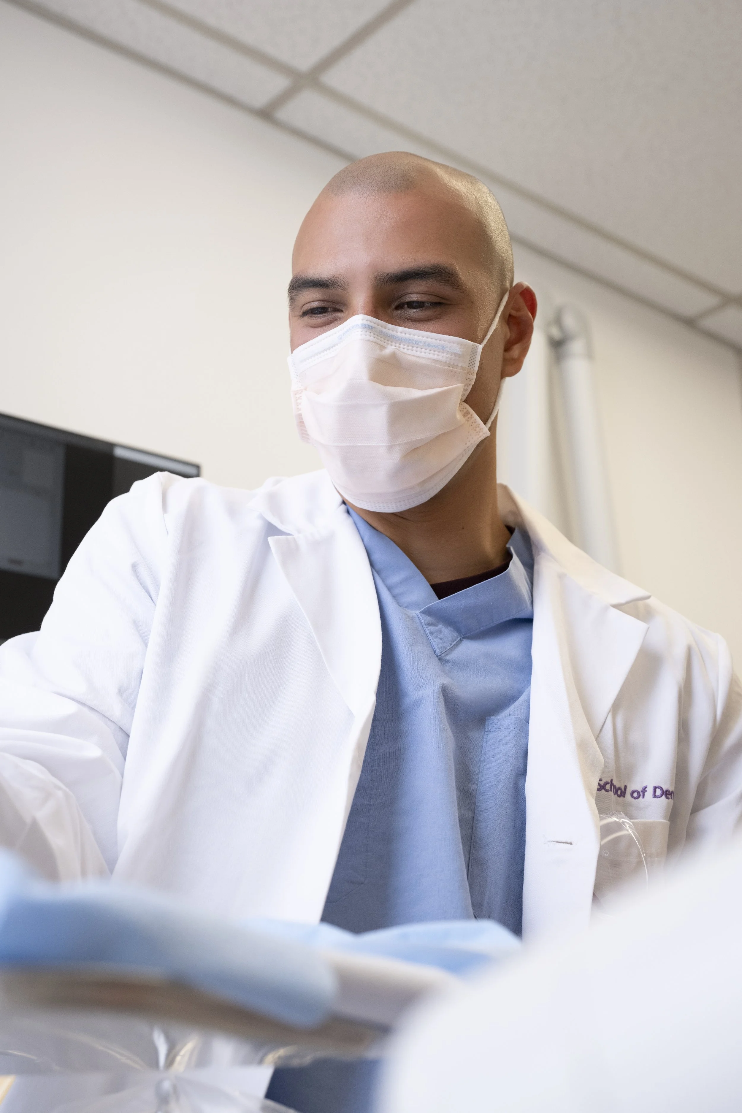 A young male healthcare professional in scrubs and a white lab coat wearing a face mask, working in a clinical setting.