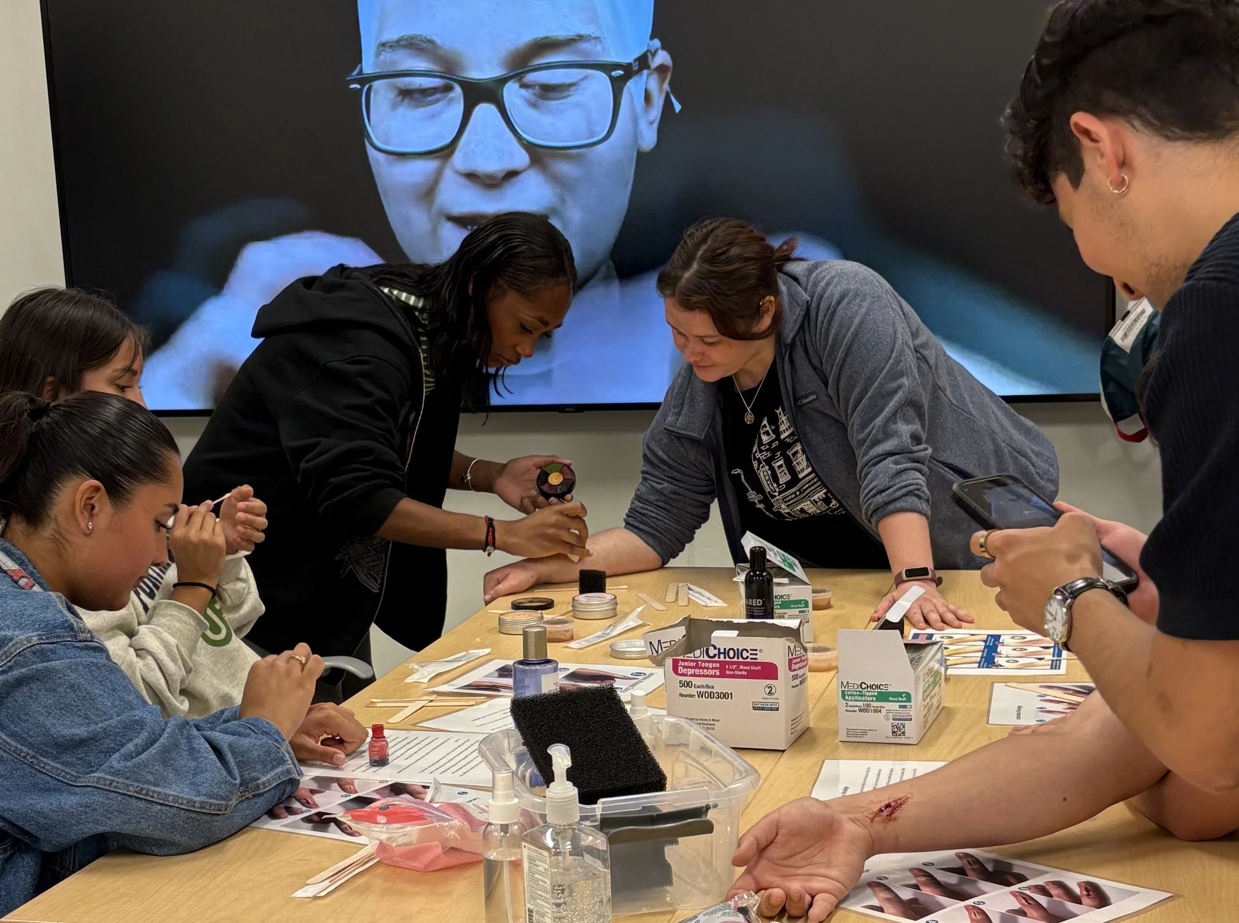 A group of people participating in a makeup or skincare workshop around a table, with a large screen displaying a close-up of a person's face with glasses in the background.