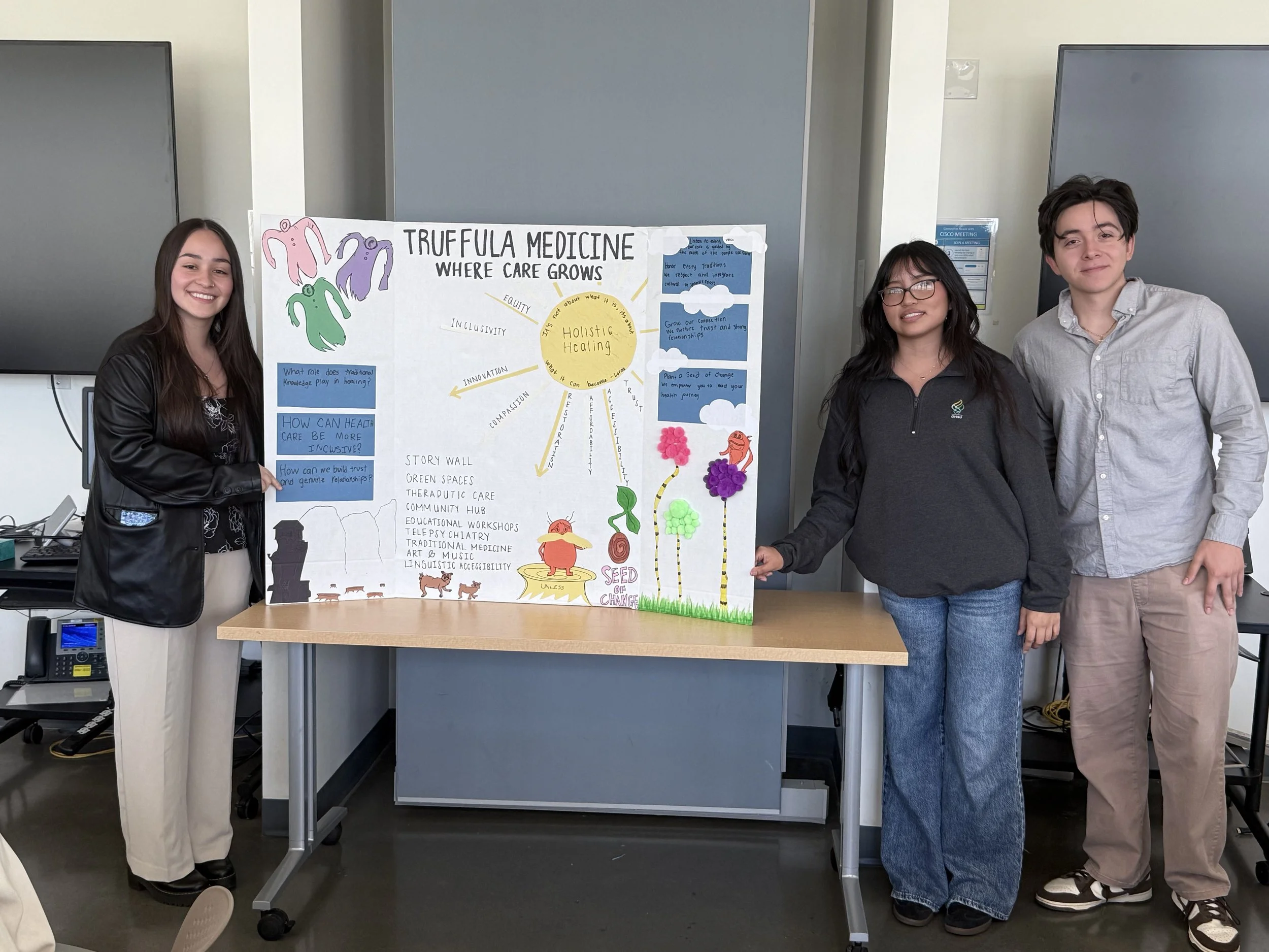 Three individuals standing next to a colorful science project display board titled 'TRUFFULA MEDICINE WHERE CARE GROWS' on a desk in a classroom or conference room.