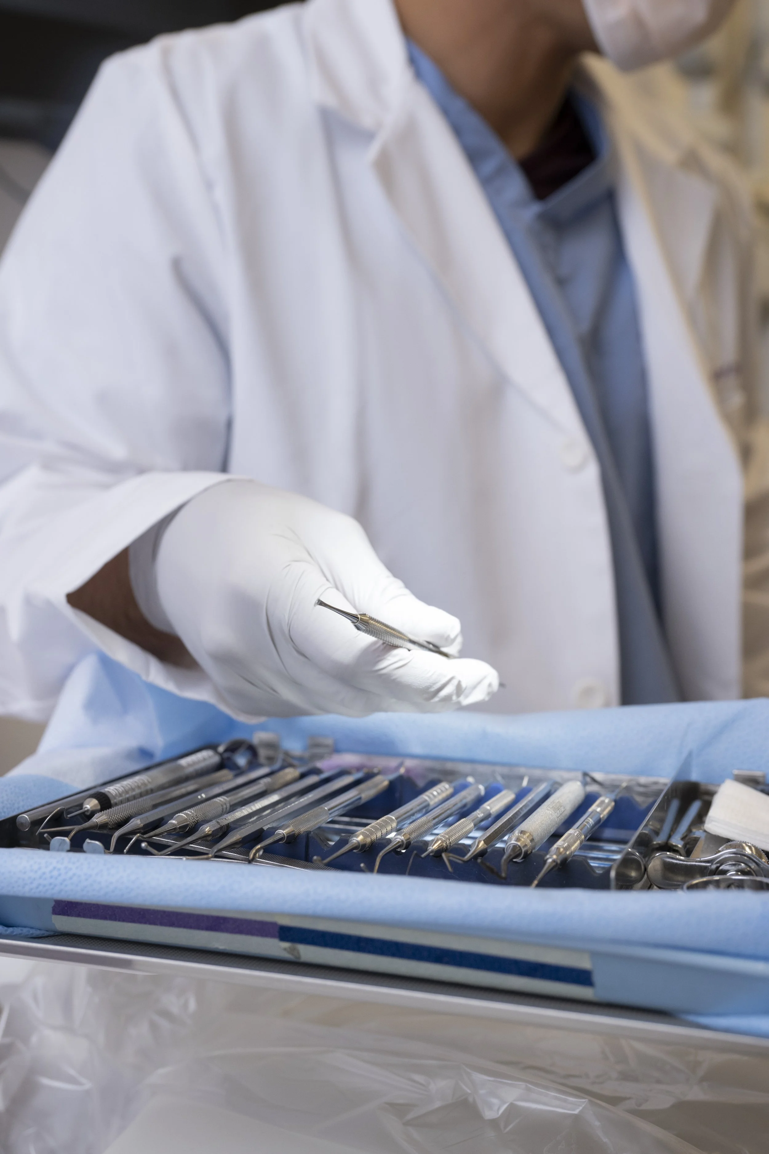 Close-up of a healthcare professional wearing a white lab coat and gloves, holding a dental instrument, with dental tools on a tray in the foreground.