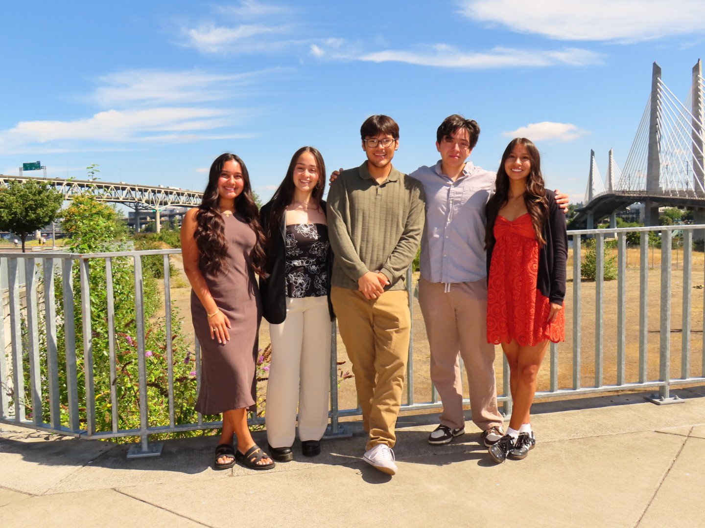 Group of five young adults posing outdoors near a railing with a bridge and blue sky in the background.