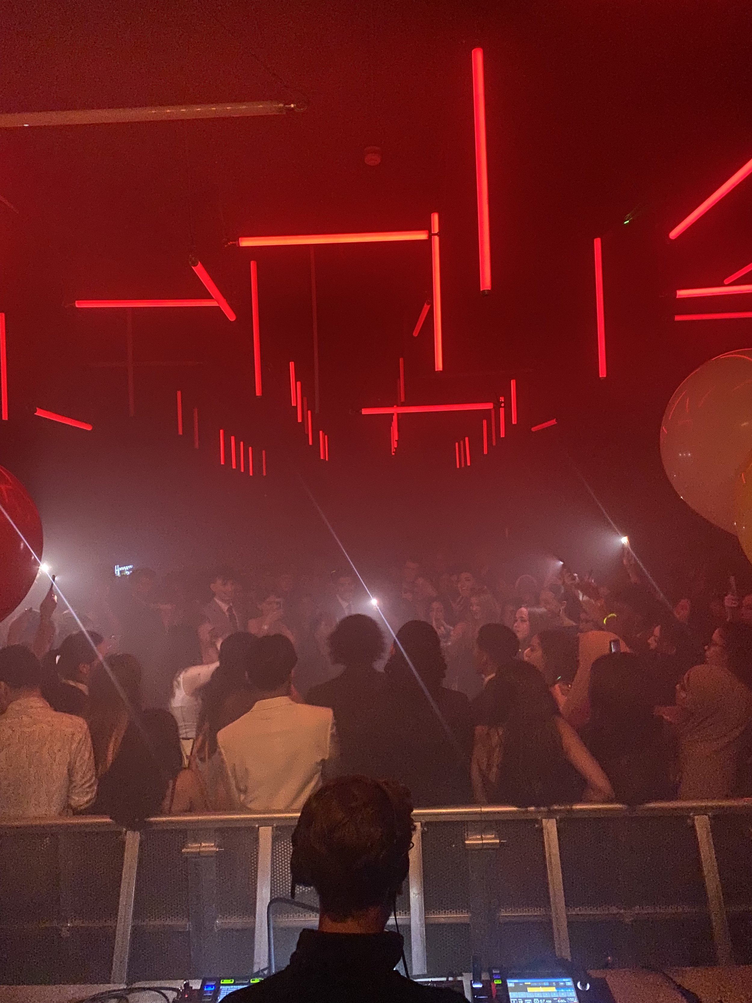Crowd of people dancing and socializing at a nightclub with red neon lights on the ceiling and DJ booth in the foreground.