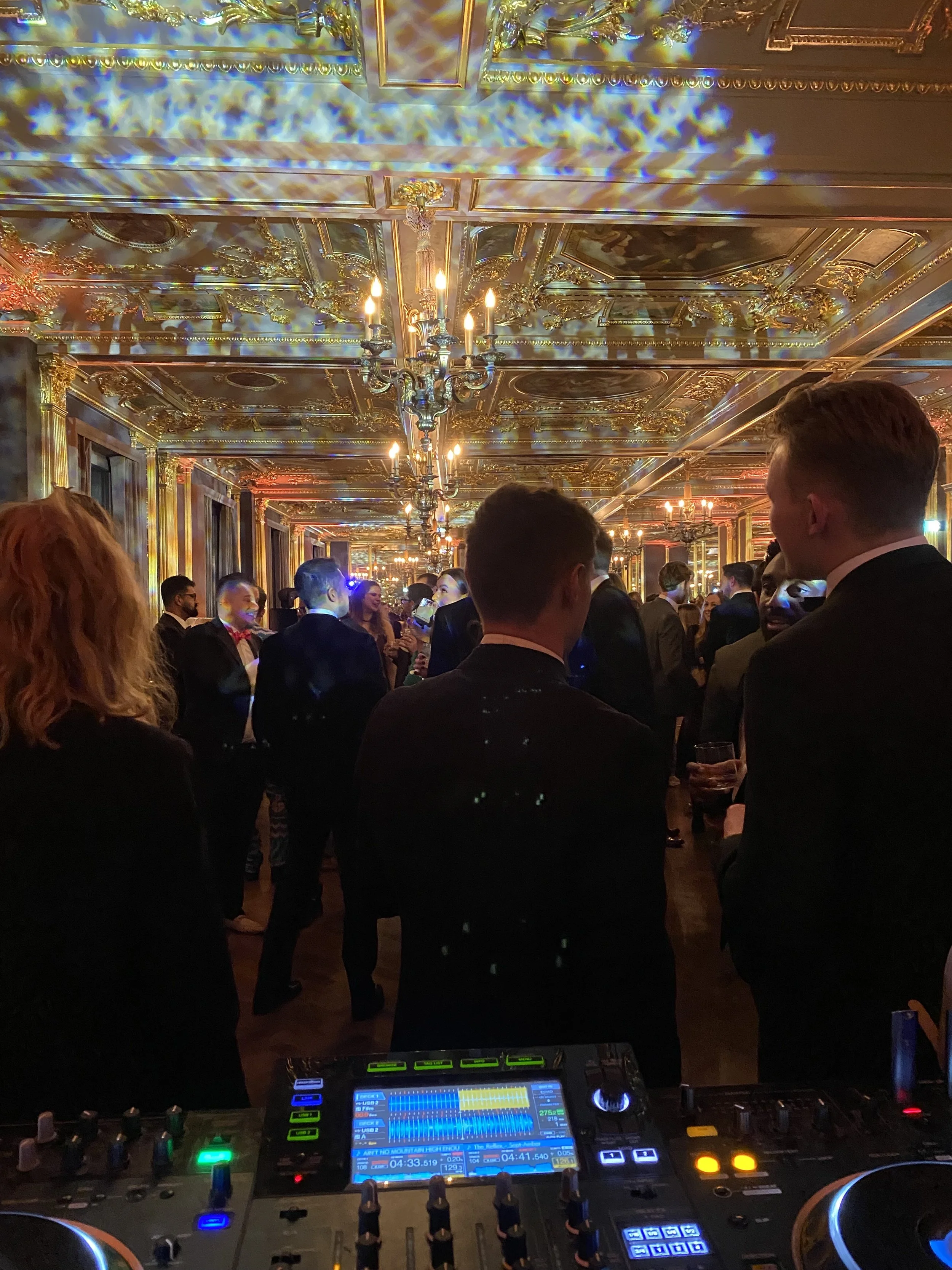 People in formal attire socializing at a fancy event with ornate gold and mirrored ceiling and a DJ setup in the foreground.
